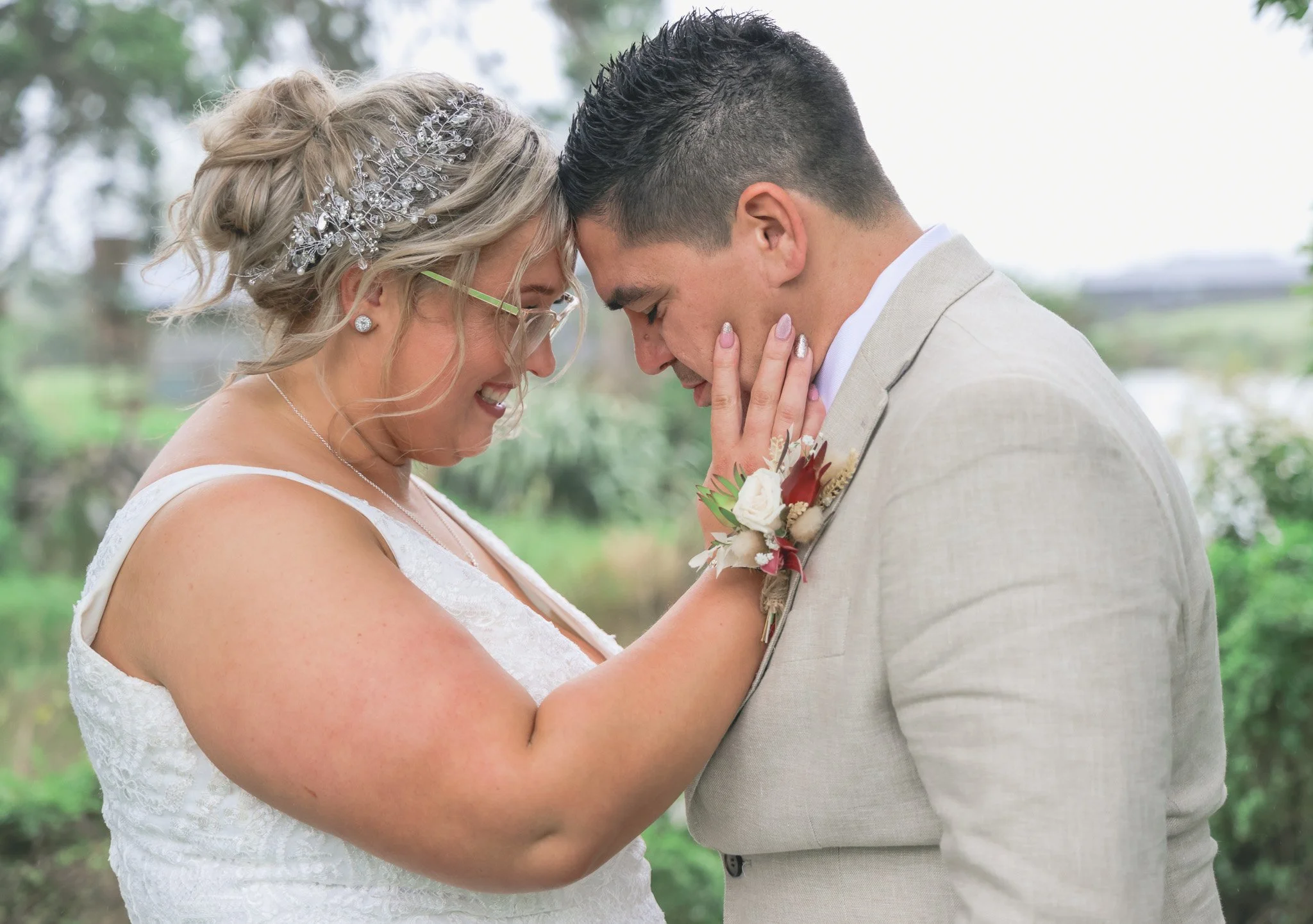 A bride and groom sharing a tender moment with foreheads touching and eyes closed outdoors, the bride wearing a white lace dress and a jeweled hair accessory, and the groom in a light-colored suit with a boutonniere.