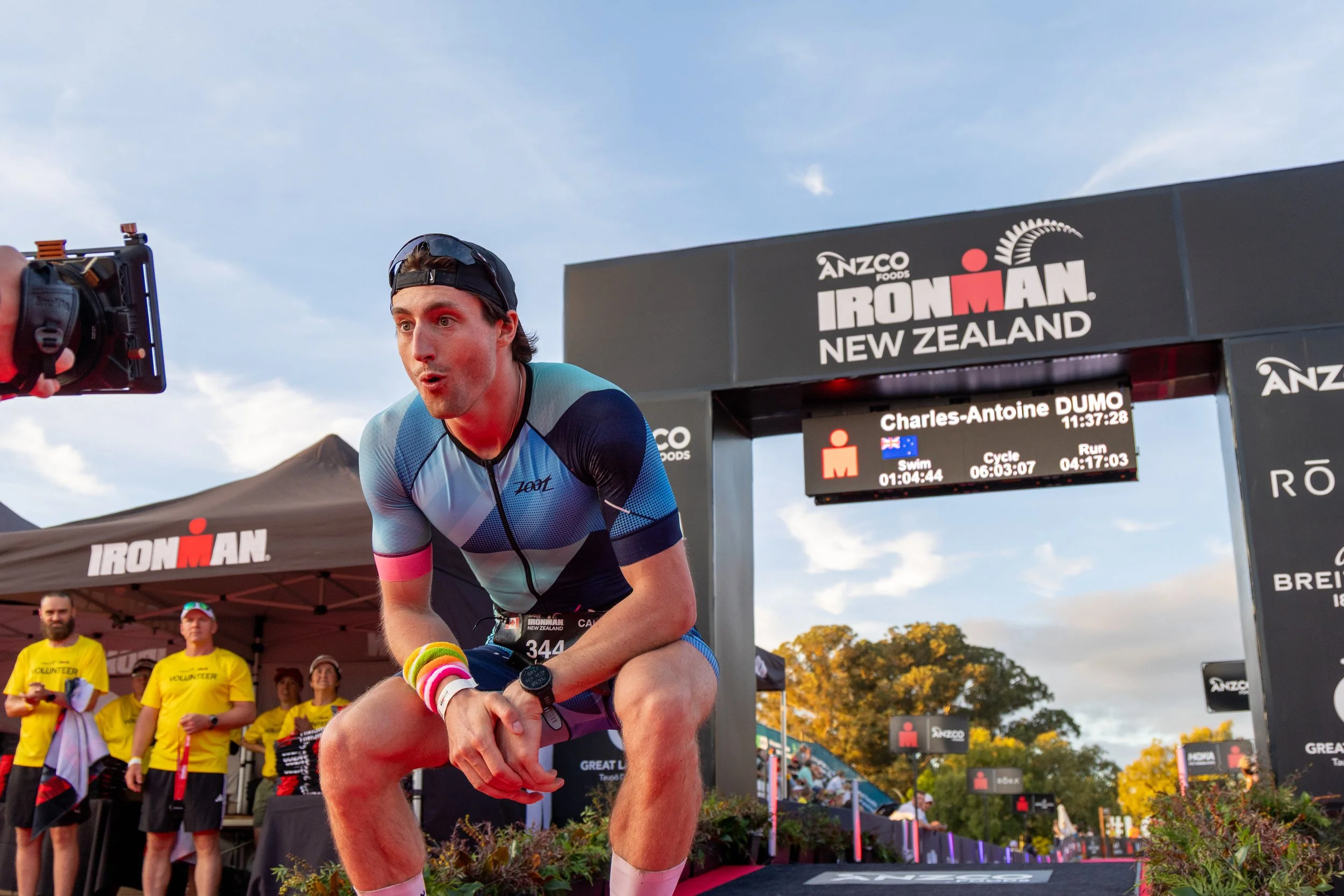 A male athlete at the finish line of an Ironman race in New Zealand, with a timing clock showing his race time. He is wearing a blue and black athletic outfit, a black cap, and colorful wristbands. Several volunteers in yellow shirts are in the backg
