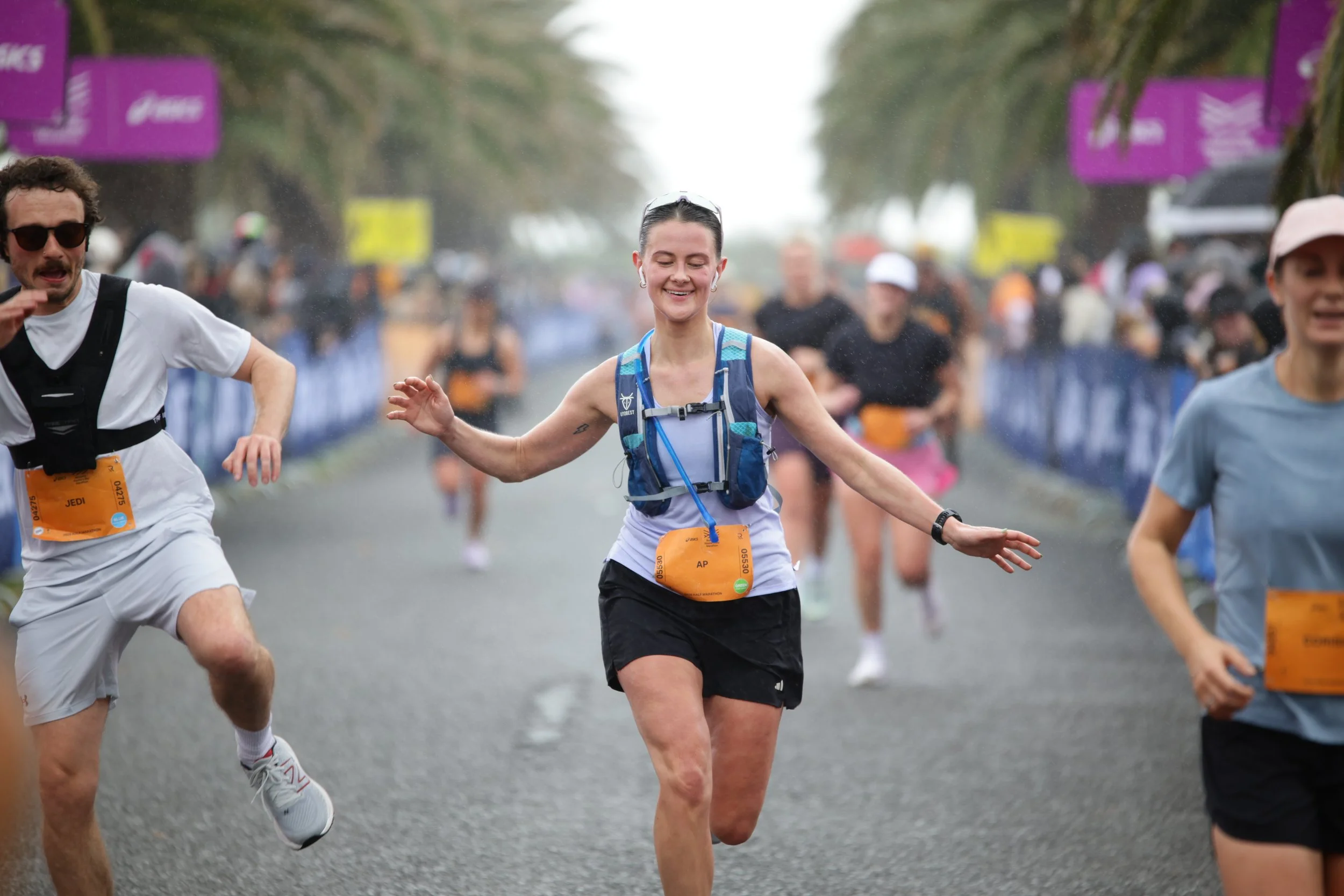 People participating in a marathon race, running on a street with trees and banners in the background, some wearing race bibs and athletic gear.