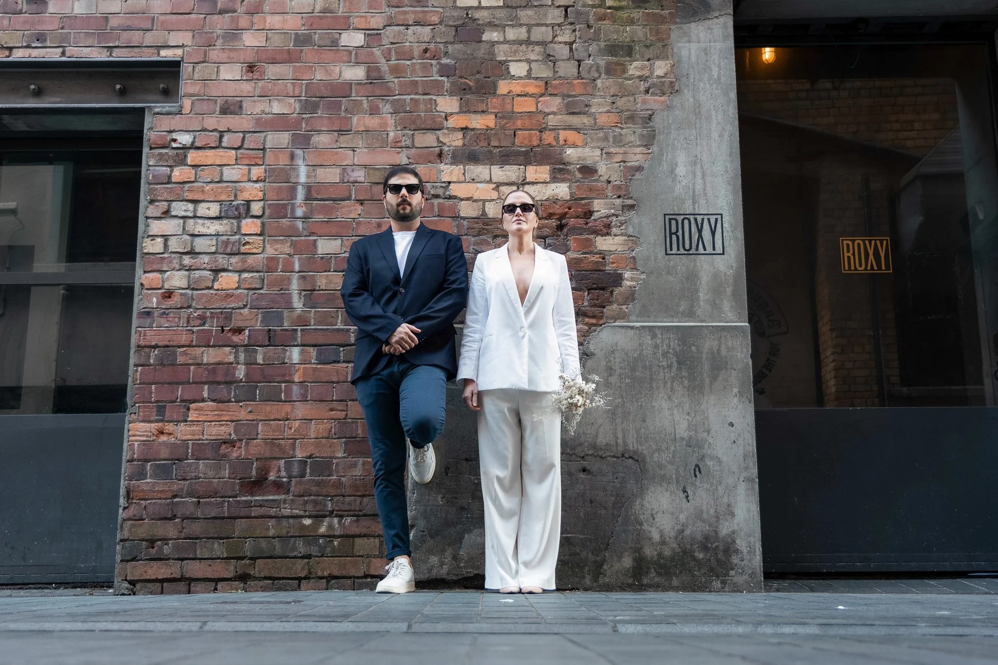 A man and woman standing against a brick wall outside a building with a sign that says 'ROXX'. The man wears a dark suit, white t-shirt, sunglasses, and has a beard. The woman wears a white suit, sunglasses, and holds a bouquet of flowers. They both 