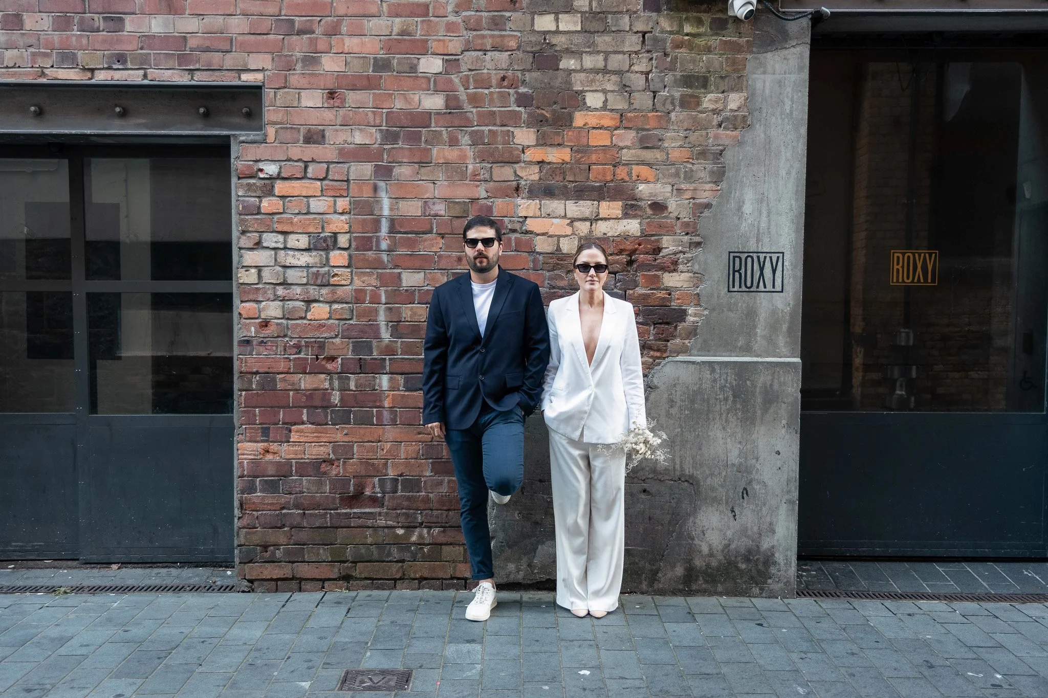 A man and woman standing against a brick and concrete wall. The man is wearing sunglasses and a navy blazer with white shirt, and the woman is dressed in a white suit holding a small bouquet of dried flowers. Both are standing outdoors on a city side