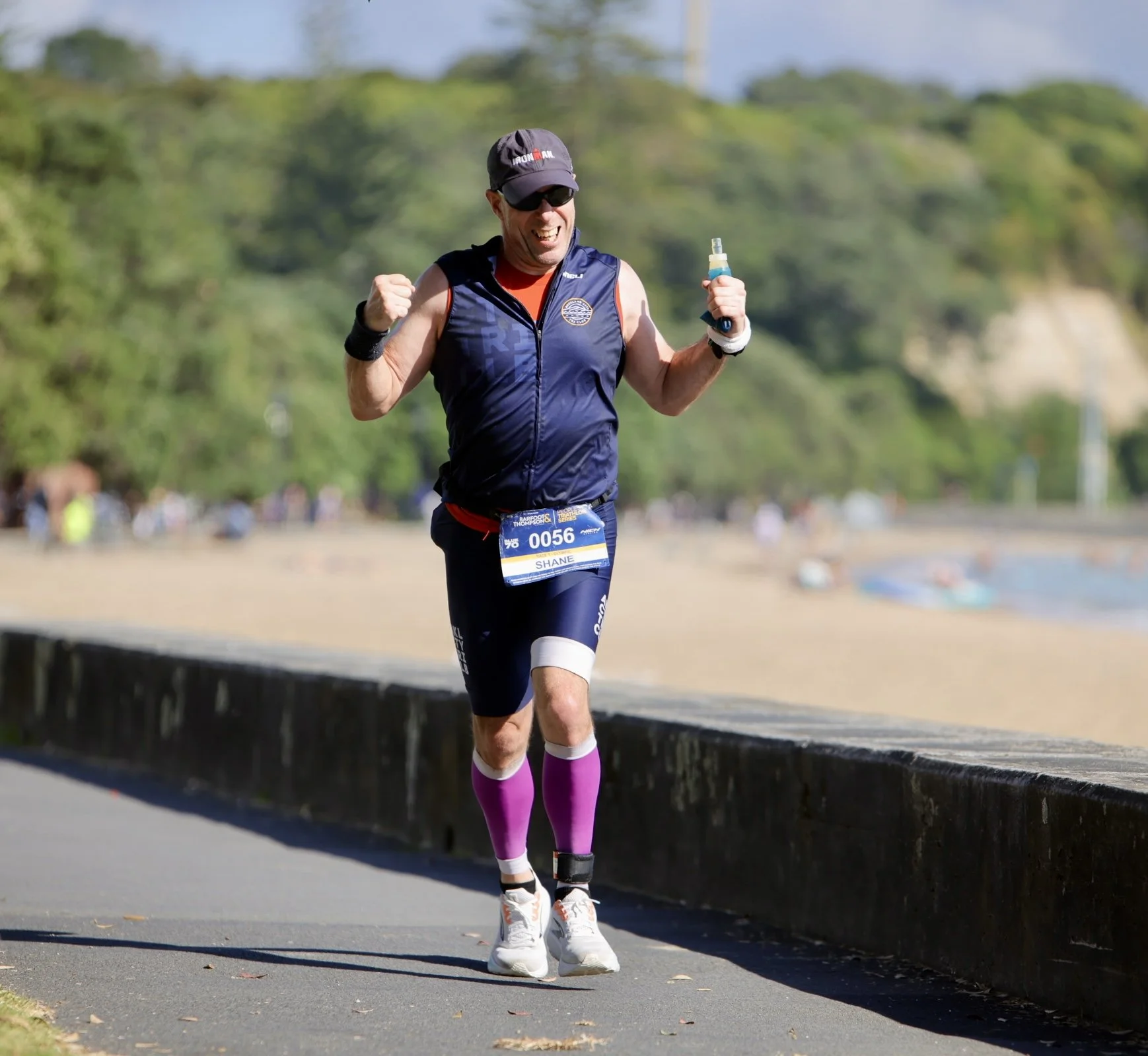 Man running outdoors during a race, wearing sunglasses, a cap, a sleeveless vest, a race bib, compression socks, and athletic shoes, smiling with fists clenched in excitement.