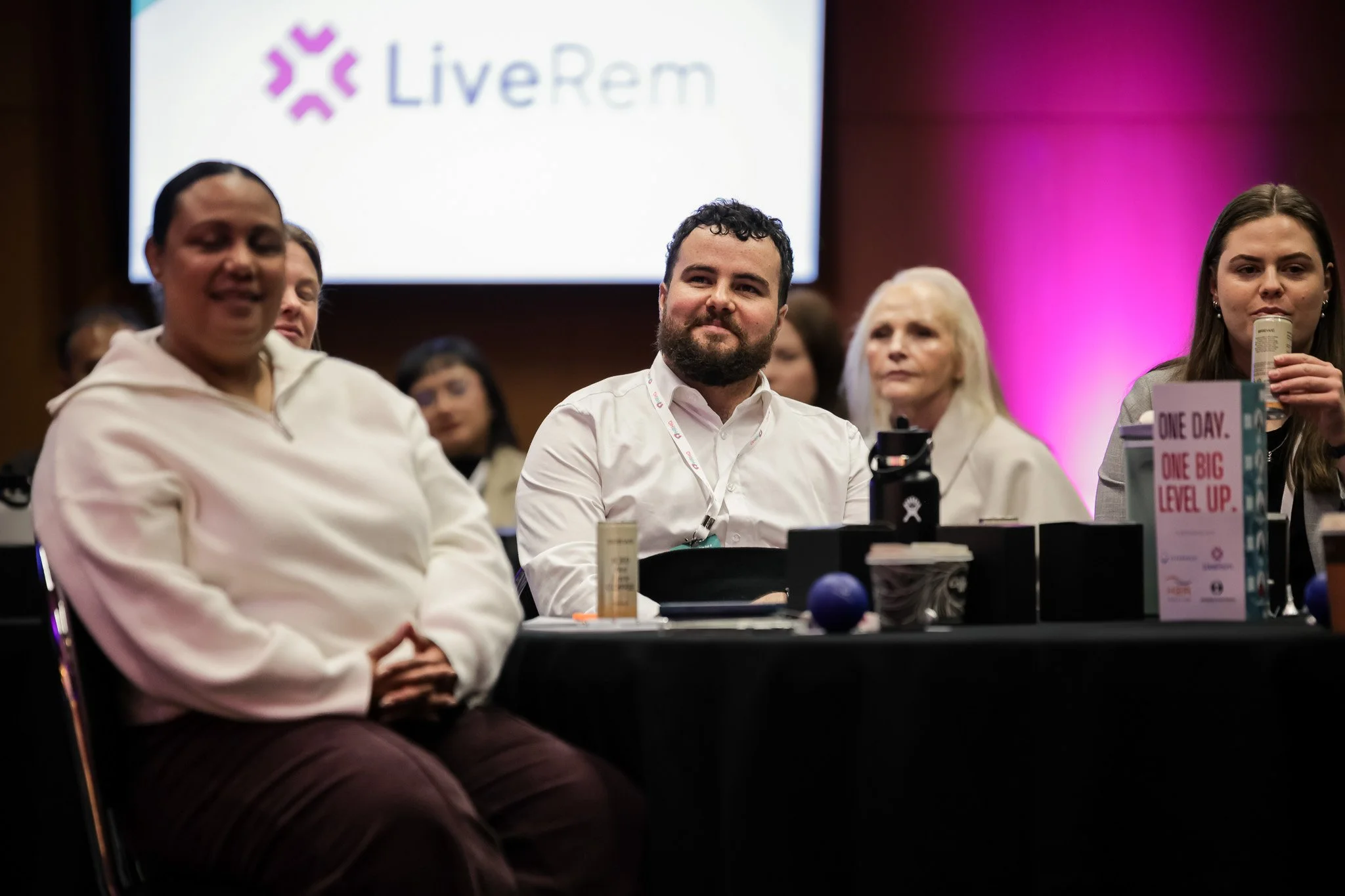 People sitting at a table during a conference or panel discussion, with a large screen behind them displaying the LivRem logo.
