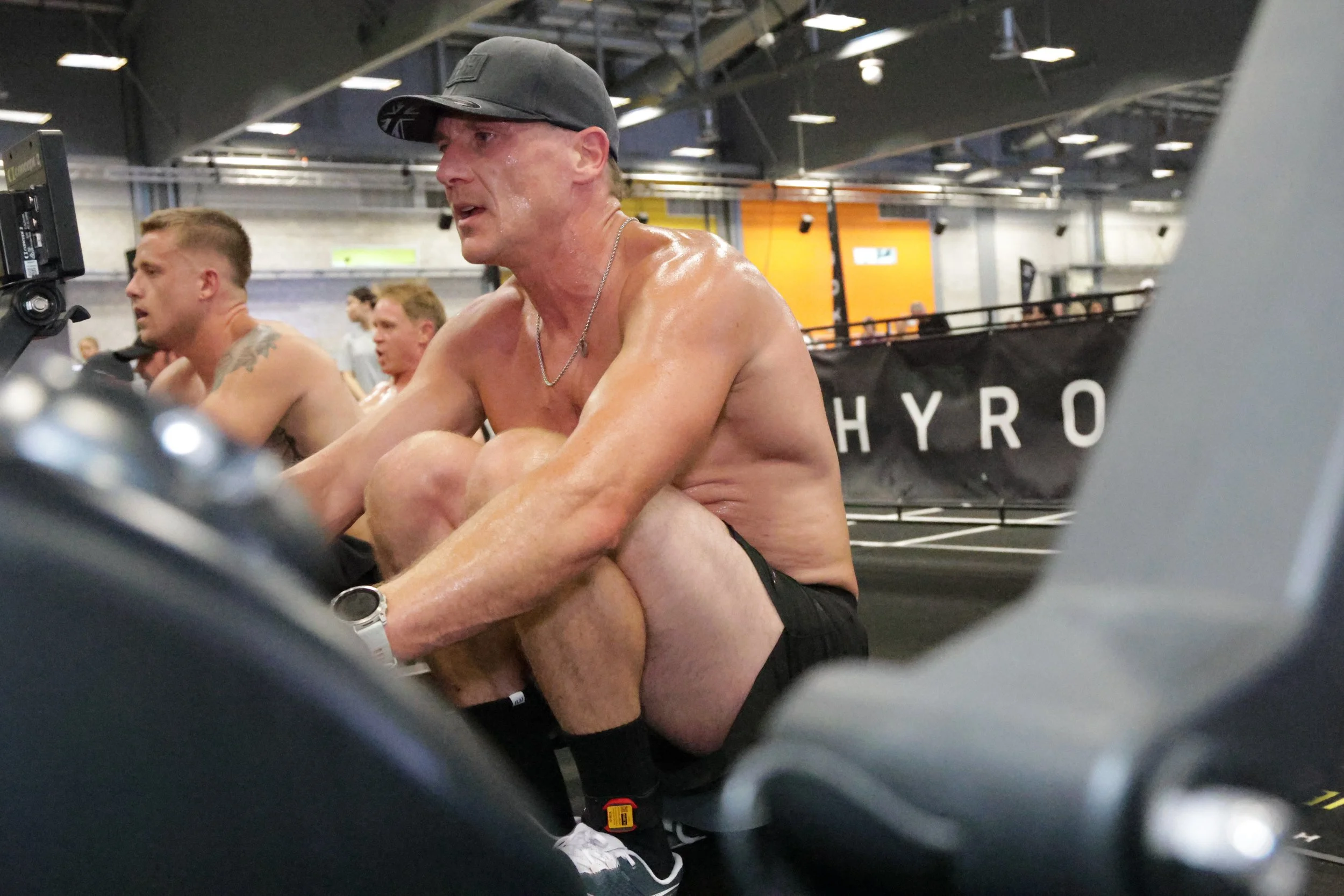 A shirtless man wearing a black cap and a silver chain, sweating and resting while exercising at a gym, with other people working out in the background.
