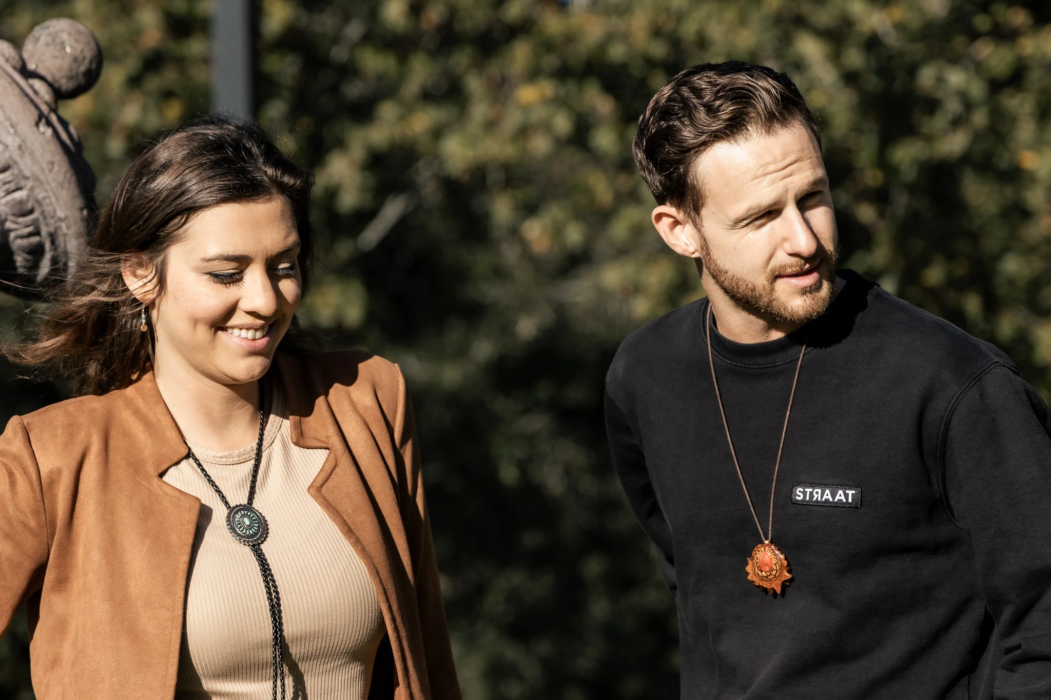 A woman and a man standing outdoors, smiling and looking down, with trees in the background.
