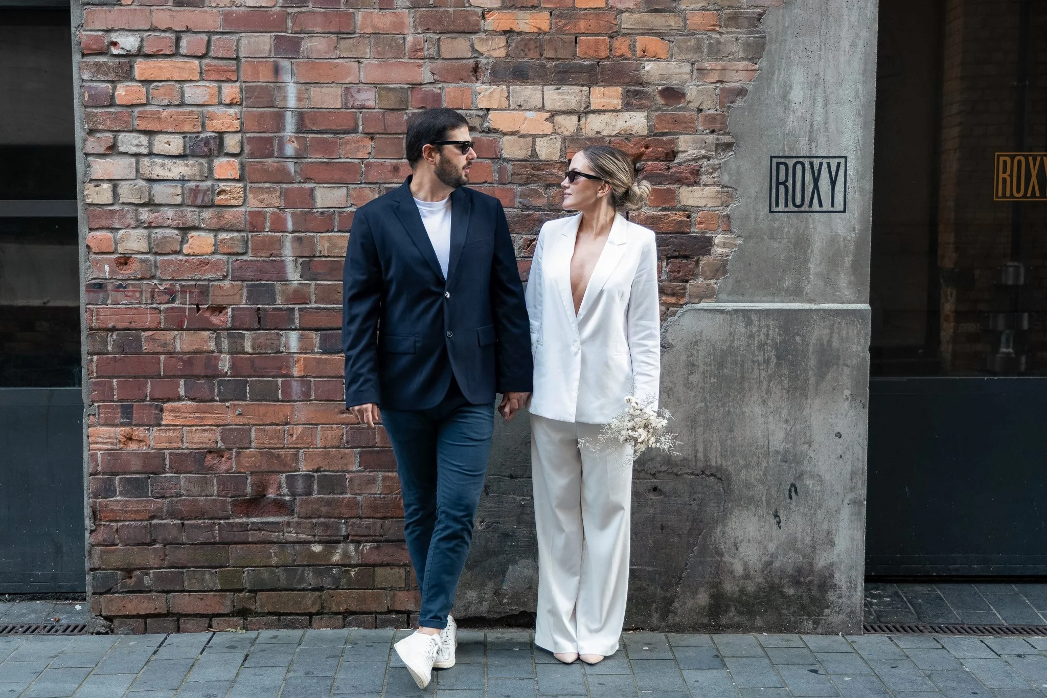 A man and woman holding hands and standing in front of a brick wall. The man is dressed in a blue suit with a white shirt and sunglasses, while the woman is wearing a white blazer and pants with sunglasses, holding a small bouquet of flowers, and loo