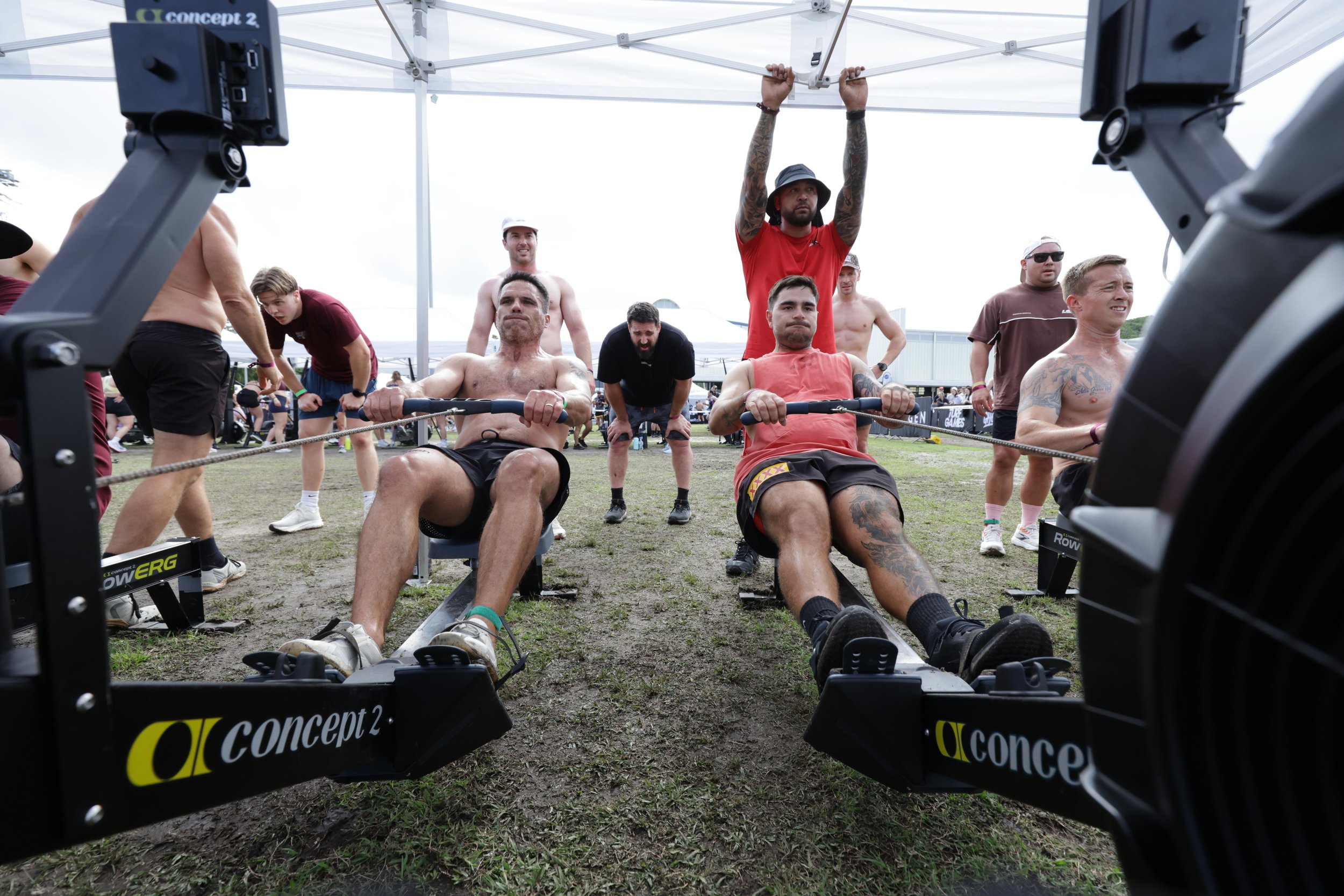 A group of people participating in an outdoor strength competition, with some sitting on rowing machines and others standing behind them, under a white canopy tent. The photo is taken from the front of the rowers, with visible equipment labeled 'conc
