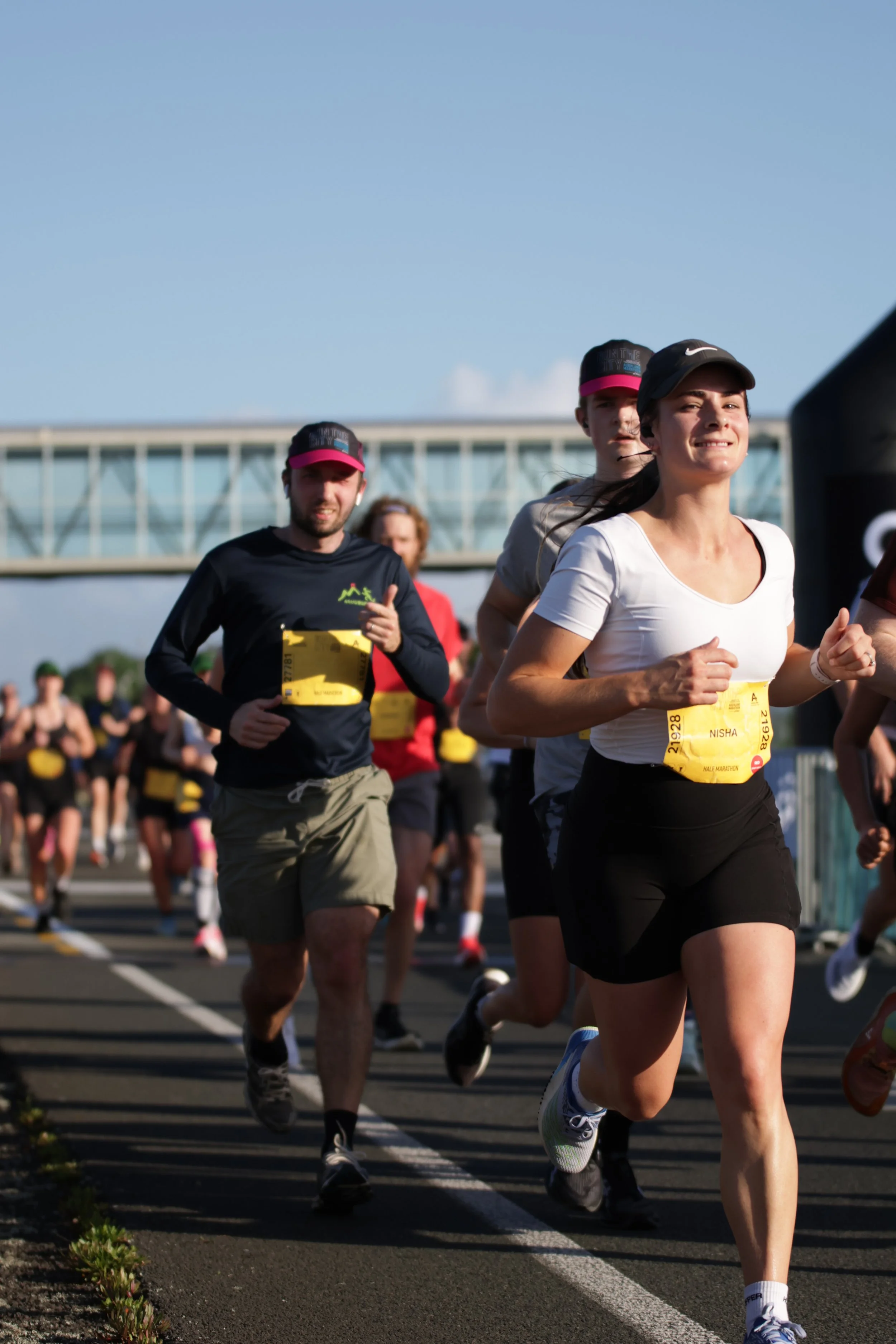 A group of runners, including women and men, participating in a marathon race outdoors on a clear day. They are running on a paved track with a building and a blue sky in the background.