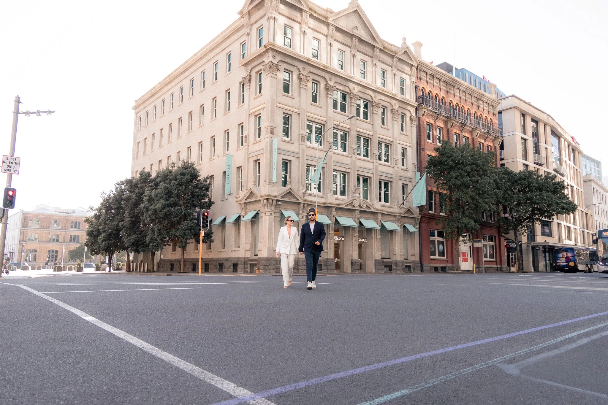 A man and woman walking across a city street intersection during daytime. The buildings surrounding them have classic architectural styles with large windows and decorative details, and there are trees and street signs visible.