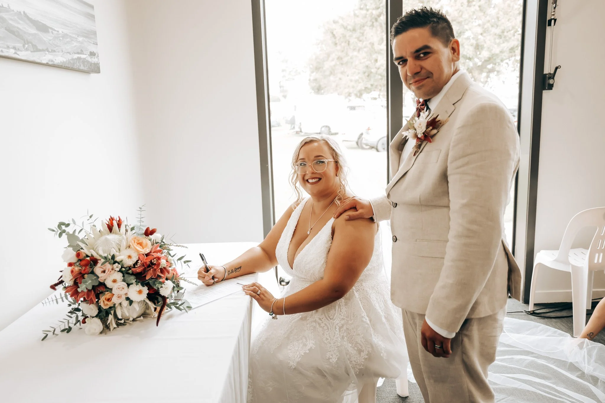 A bride in a white wedding dress signing a document, supported by a man in a light beige suit with a boutonniere, at a wedding reception with a bouquet of flowers on a white table.