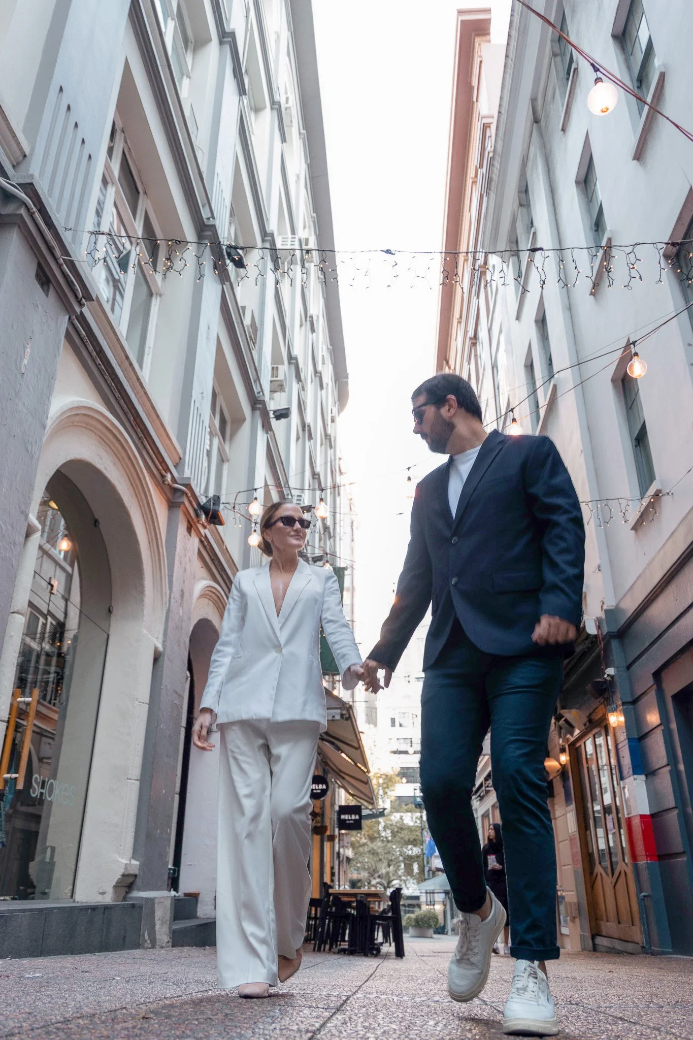 A man and a woman holding hands and walking in an urban street, with string lights hanging above, tall buildings on both sides, and shops at street level.