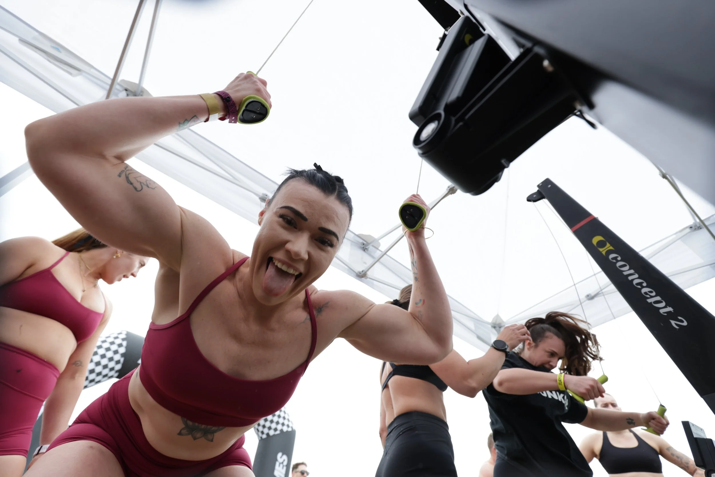 Women participating in a fitness class, lifting small green dumbbells, under a white canopy tent, with a woman smiling and sticking her tongue out in the foreground.