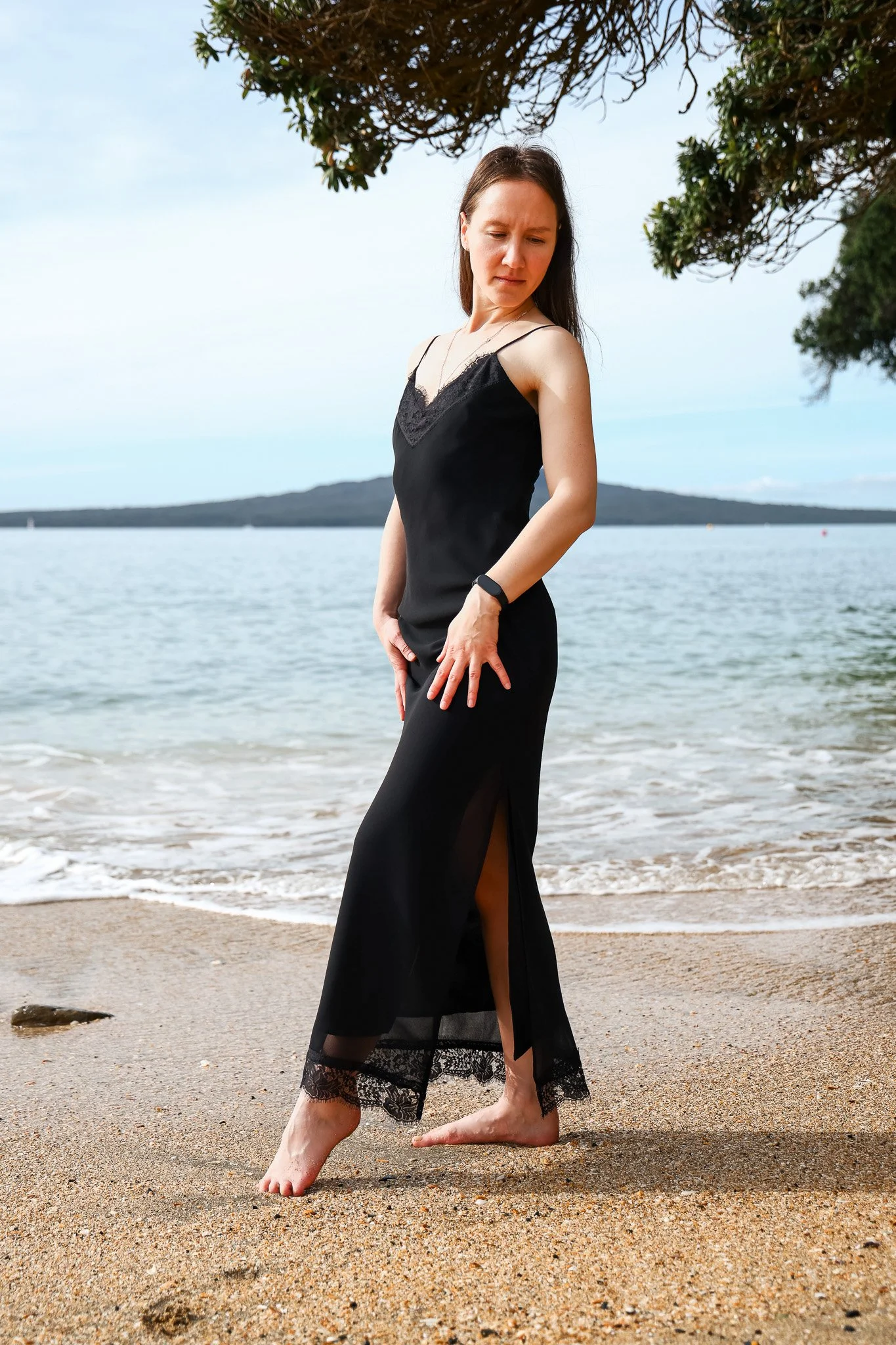 A woman in a black dress with lace details standing barefoot on a sandy beach. She is looking down and slightly to her right, with her right hand resting on her thigh and her left arm hanging by her side. There are trees overhead and the ocean in the