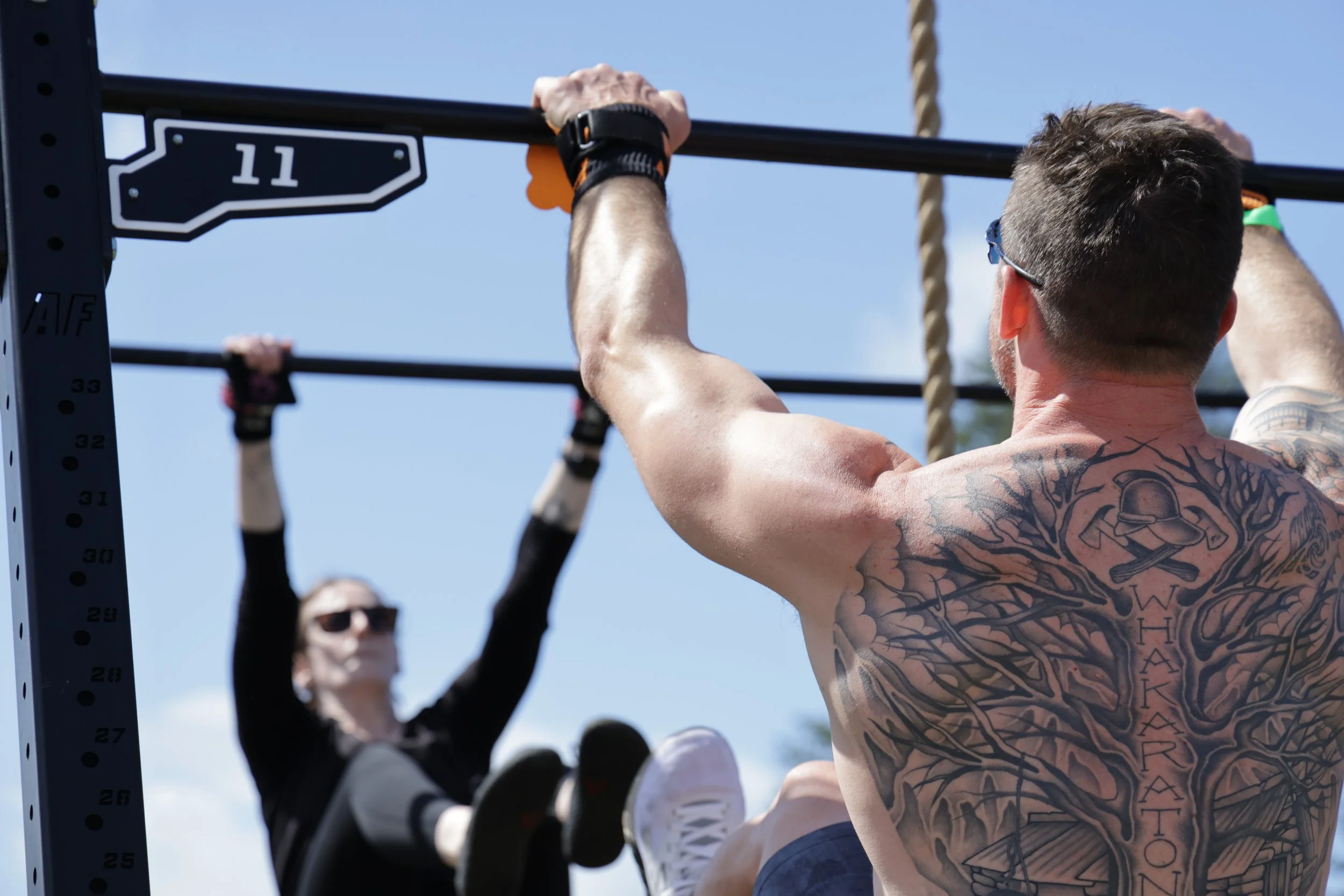 A man with tattoos on his back and arm is doing pull-ups on a black pull-up bar during outdoor exercise, with other people in the background.