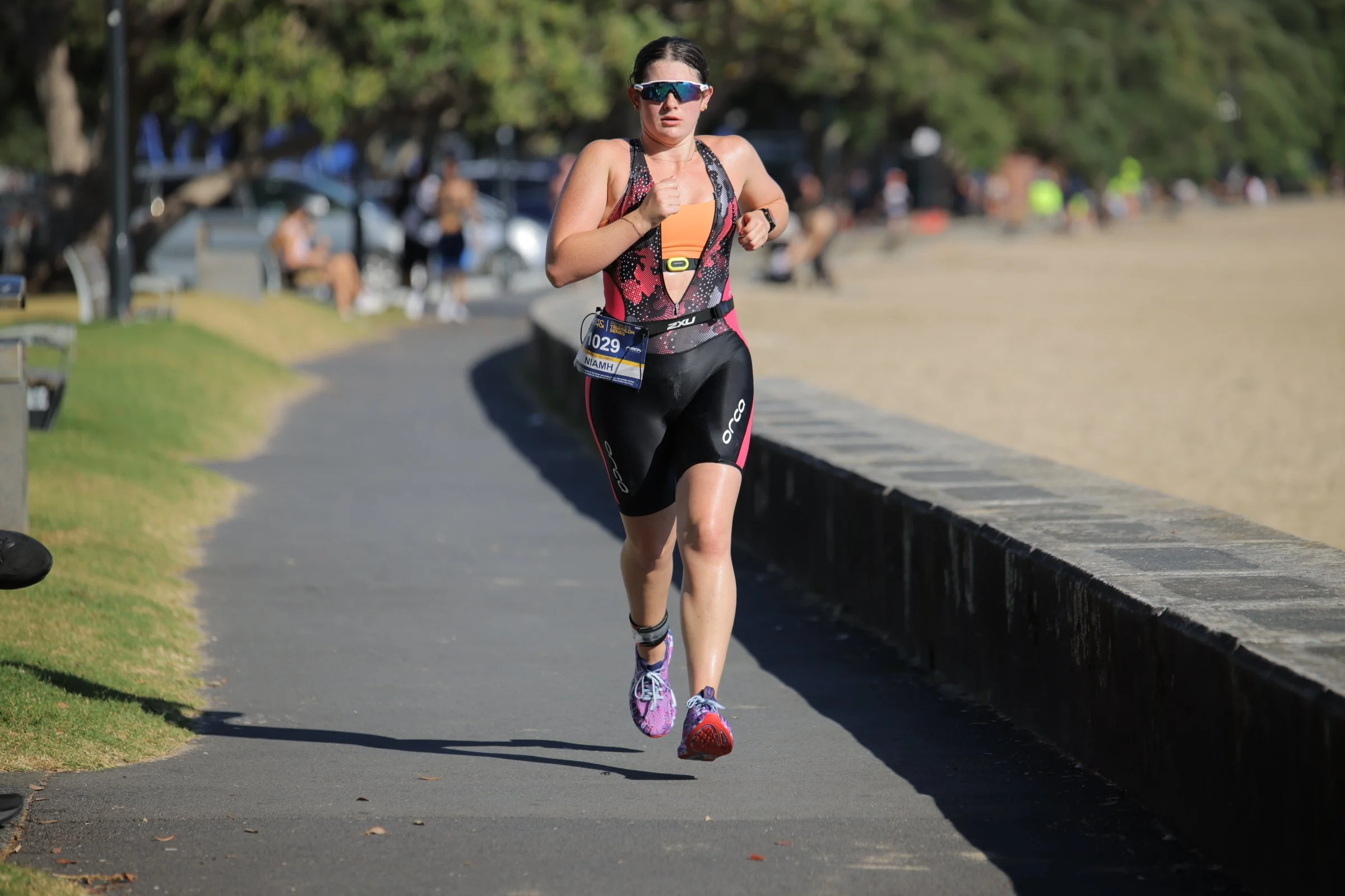 A woman running in a race, wearing sunglasses, a black and pink athletic outfit, purple running shoes, and a race bib numbered 1029.