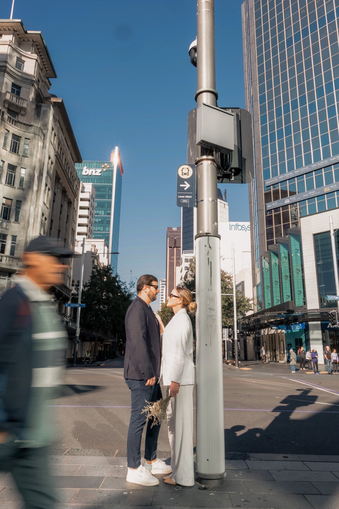 A man and woman in formal clothing standing face-to-face on a city street corner, near a pole with surveillance cameras, with tall buildings and a blue sky in the background.