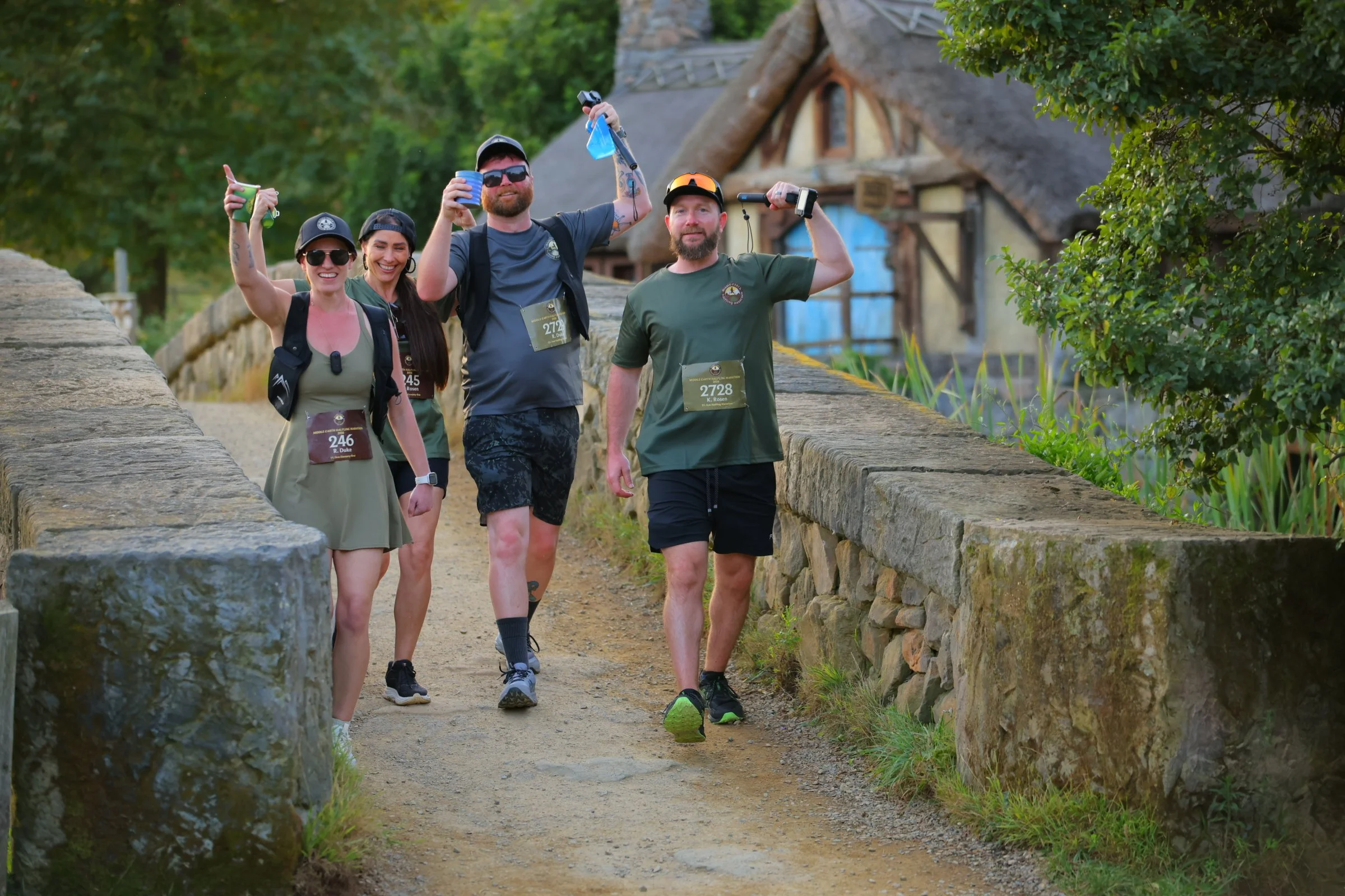 Group of five hikers walking on a stone path in a wooded area with greenery and an old cottage in the background. They are smiling and holding drinks, wearing outdoor clothing and backpacks.