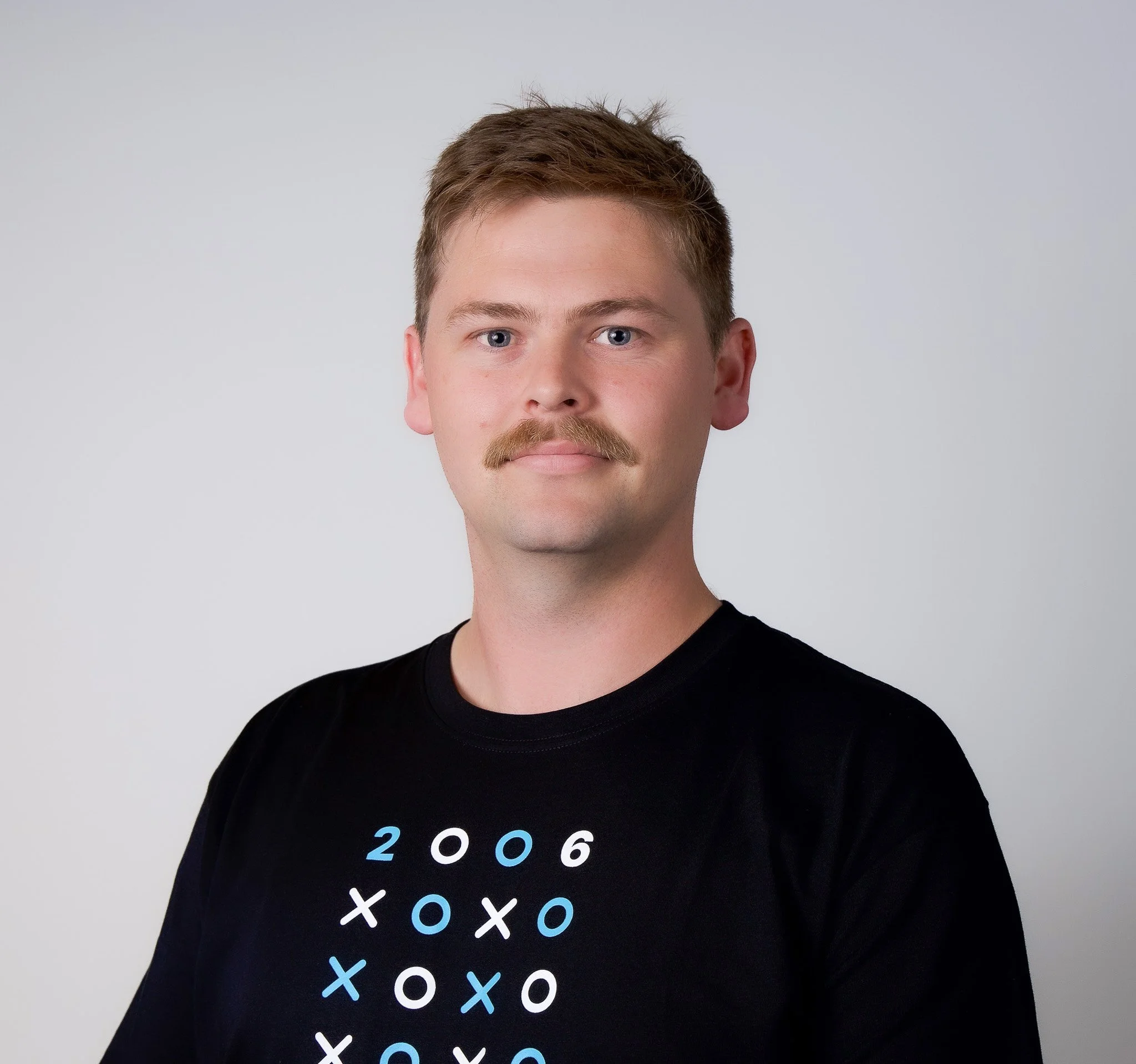 A young man with short brown hair and a mustache wearing a black T-shirt with a tic-tac-toe design and the year 2006 on it, against a plain light gray background.