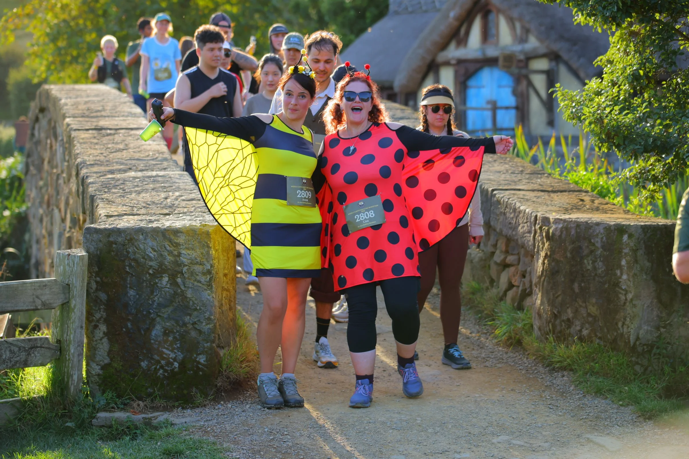 Group of people participating in a fun run or walk event on a sunny day, with two women at the front dressed in colorful, superhero-themed costumes with capes, leading the group across a stone bridge in a rural setting.