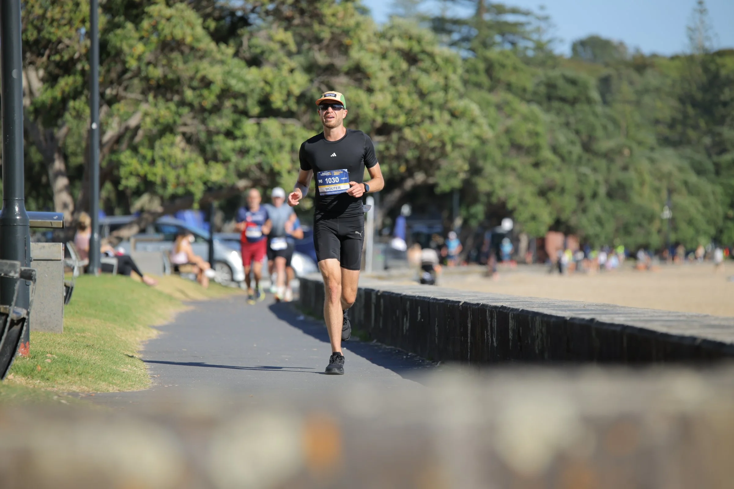 A man running in a park with a medal around his neck, wearing sunglasses, a black t-shirt, black shorts, and a cap, with trees and other people in the background.