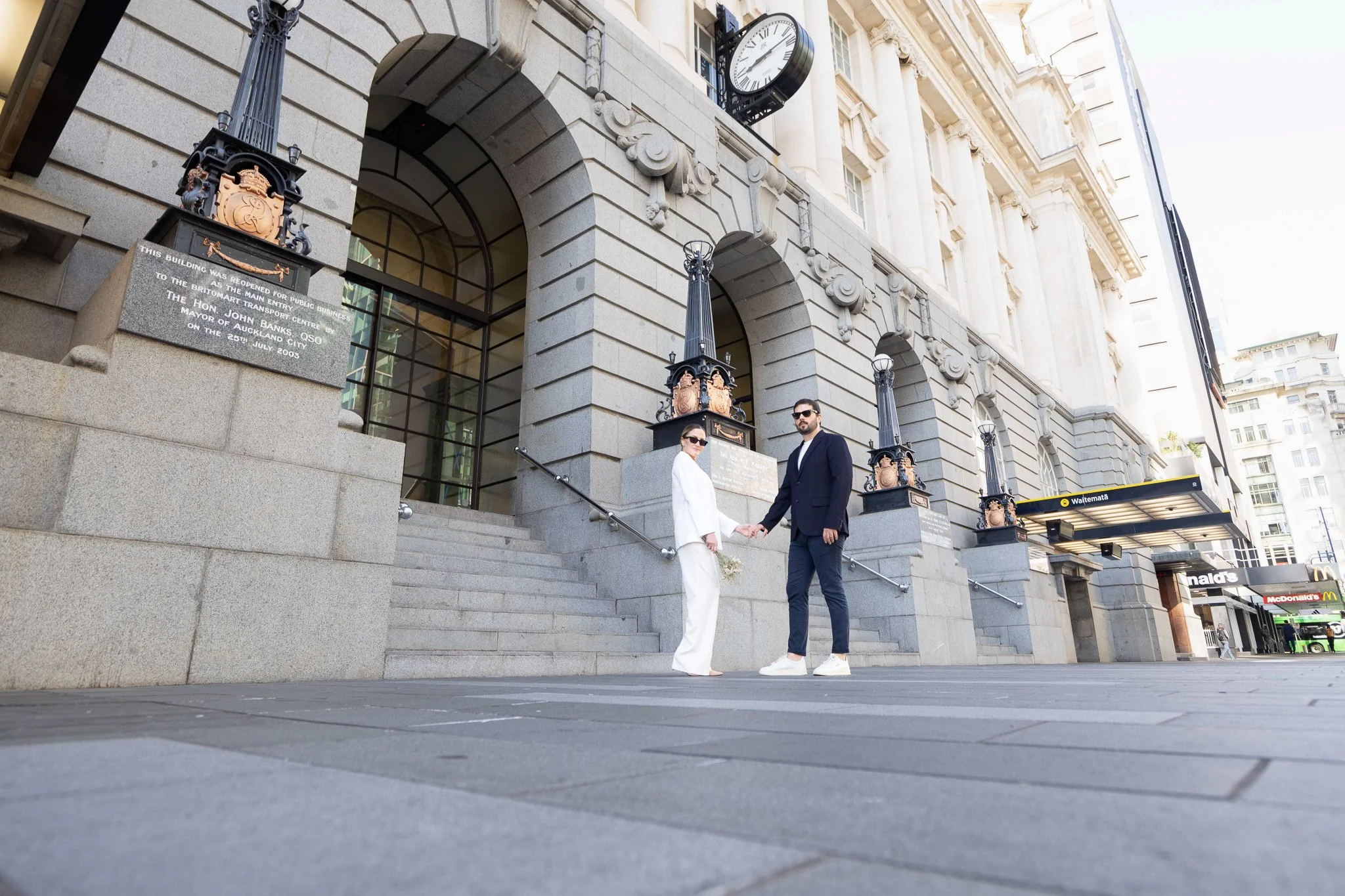 A man and woman in formal attire holding hands on the steps outside a historic building, with street and storefronts visible in the background.