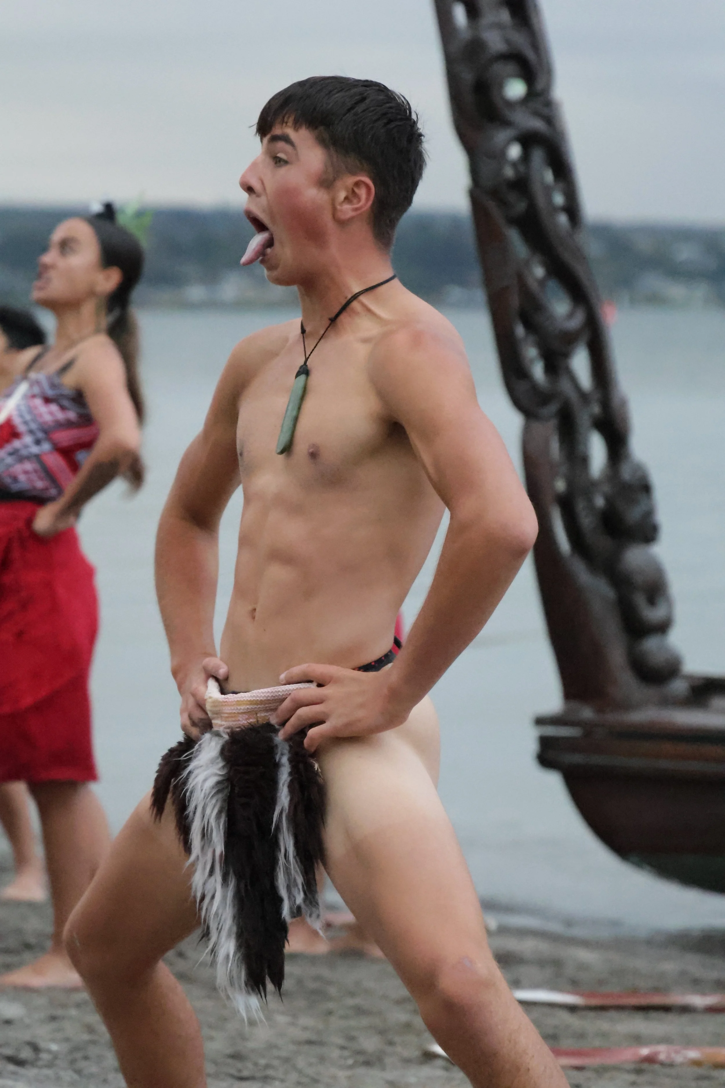 A shirtless person with short dark hair making a funny face with their tongue out and eyes wide open, standing outdoors near water with a wooden sculpture and other people in the background.