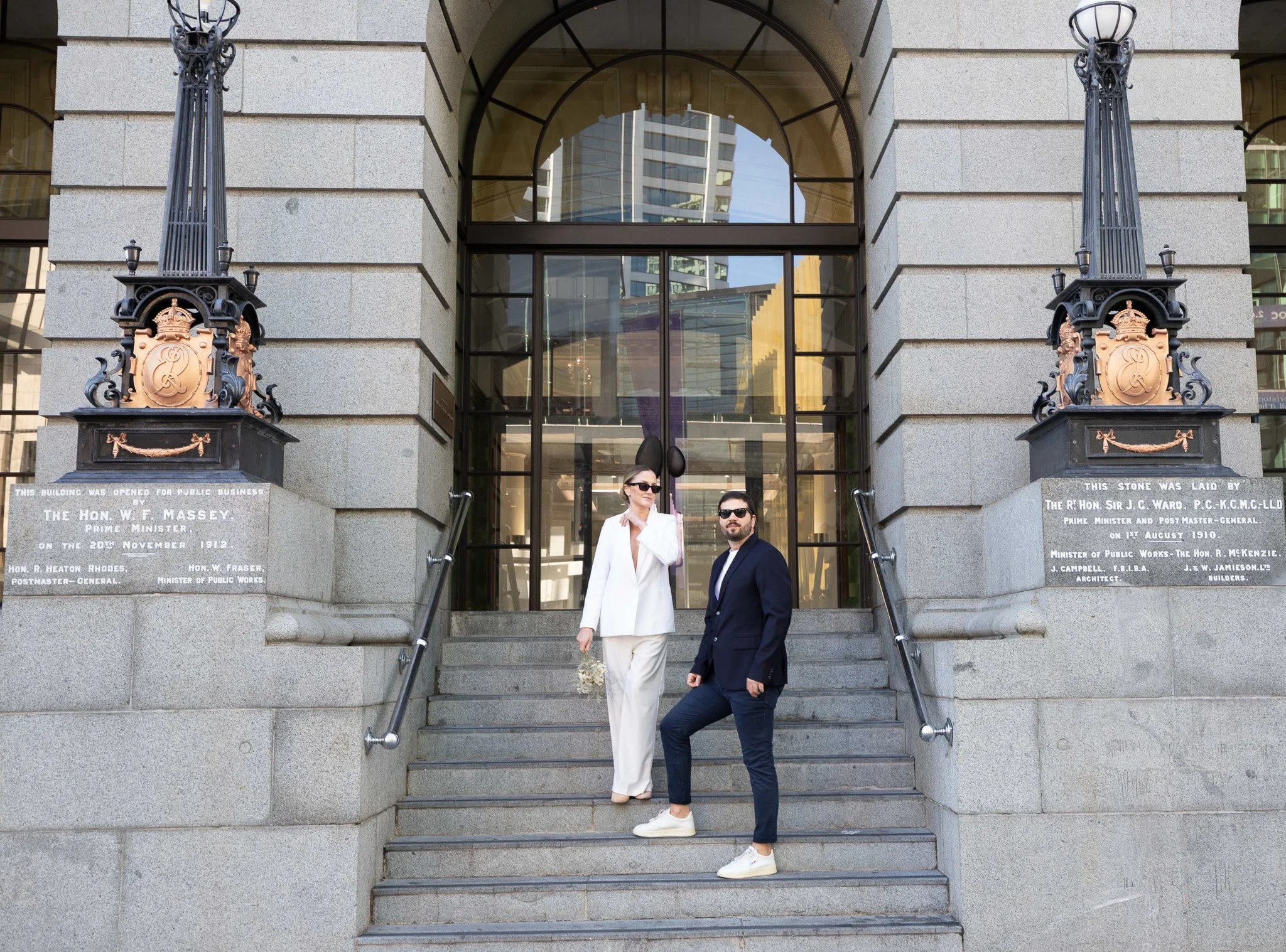 Two businesspeople, a woman in a white suit and a man in a dark suit, standing on front steps of a historic stone building with ornate lanterns and inscriptions, with modern skyscrapers reflecting in the glass behind.