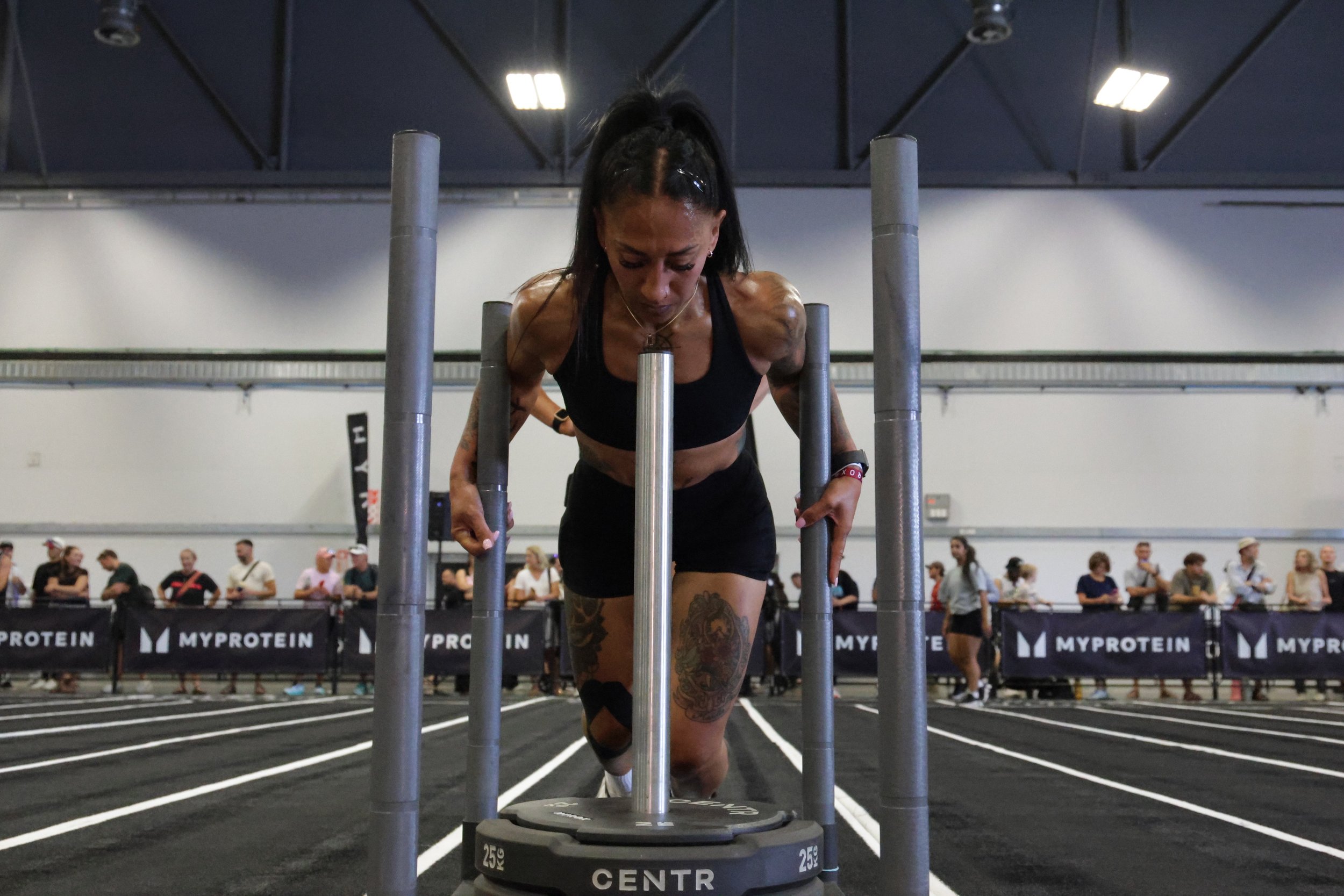 Female athlete in black sports bra and shorts performing a sled push exercise at a fitness event, with spectators in the background.