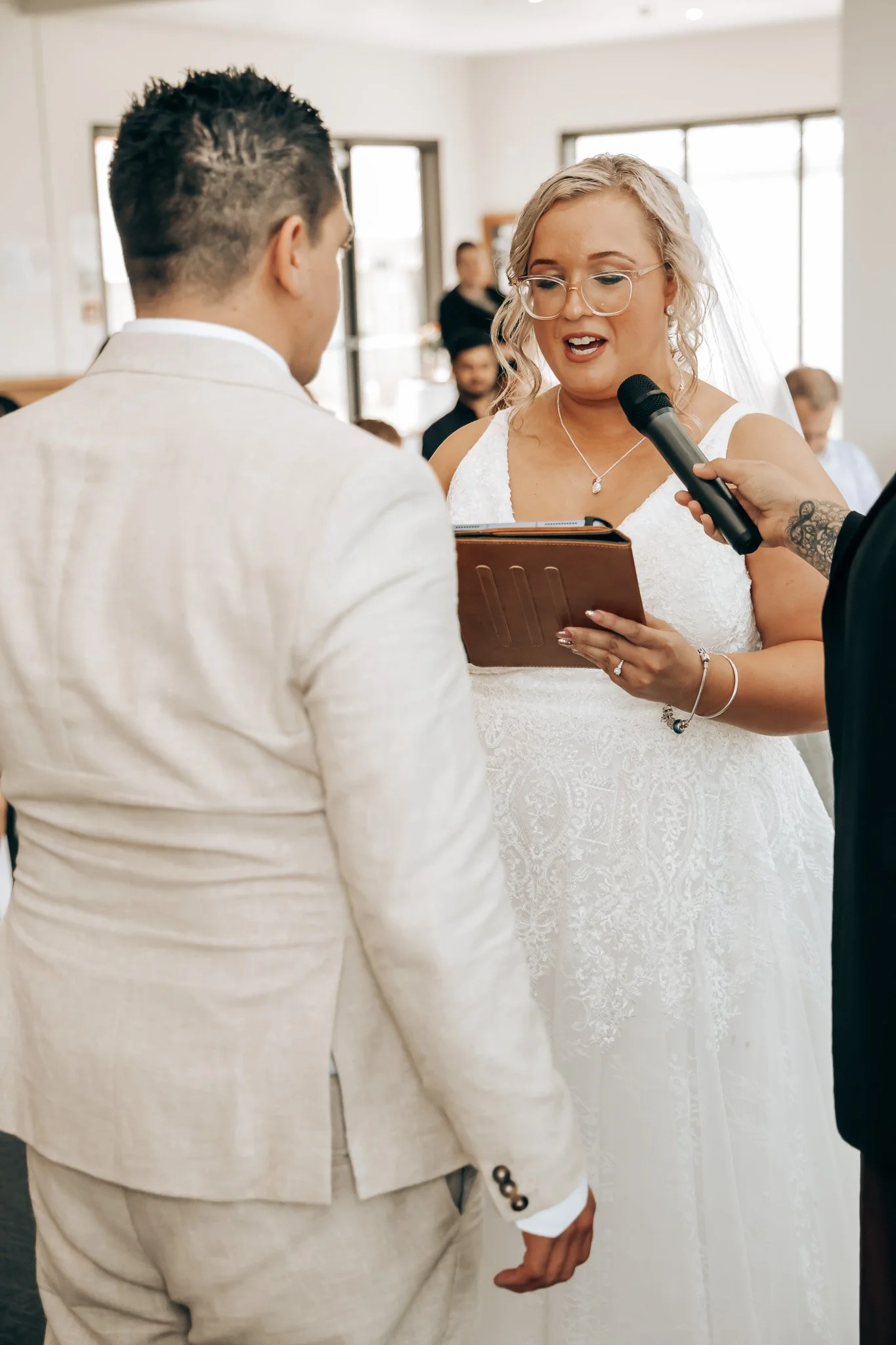 Bride and groom during wedding ceremony with officiant reading vows, bride wearing lace wedding dress and glasses, groom in beige suit.