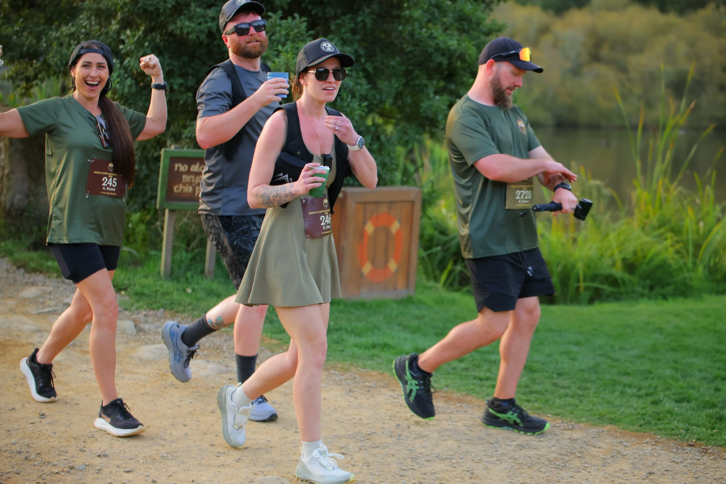 Group of people running outdoors on a trail near a body of water, some smiling and wearing athletic clothing, with greenery in the background.