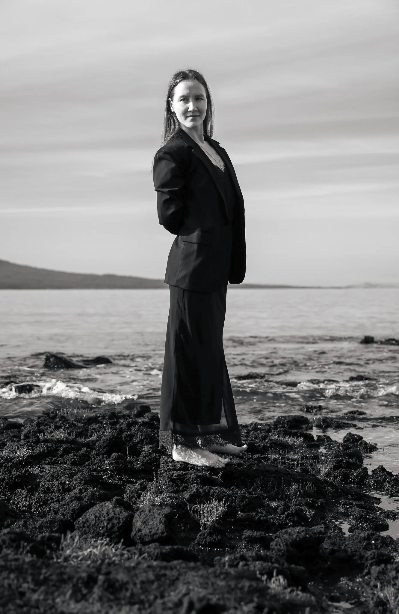 A woman in a dark blazer and skirt standing barefoot on rocks by the water, facing the camera, with a landscape of water and mountains in the background in black and white.