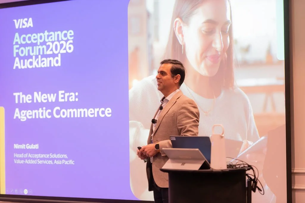 A man in a tan blazer giving a presentation on stage at a conference, with a large screen behind him displaying the title 'Acceptance Forum 2026 Auckland' and the topic 'The New Era: Agentic Commerce.' The screen also shows a picture of a woman with 
