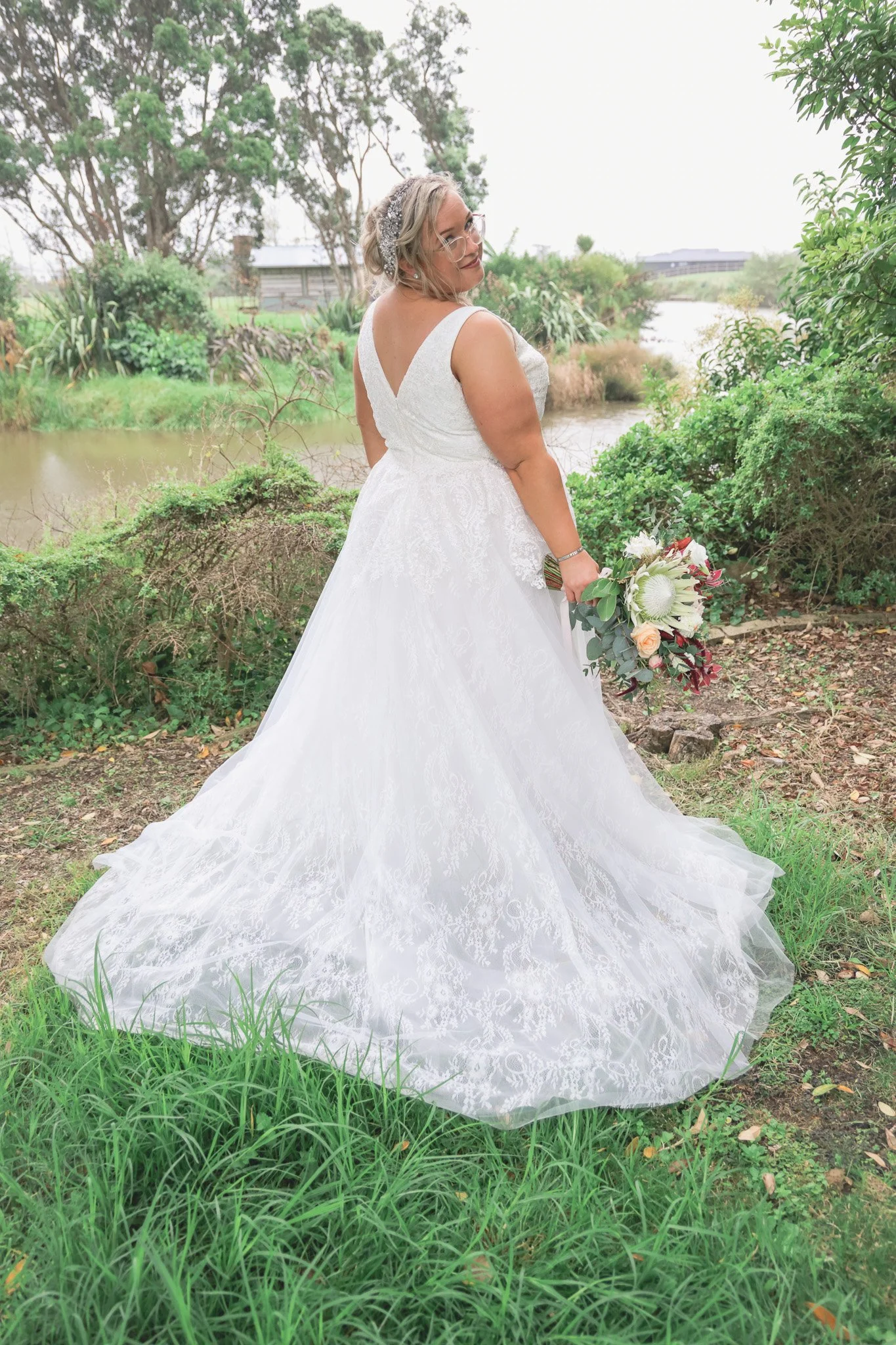 A woman in a white wedding dress holding a bouquet outdoors near a body of water, with trees and greenery in the background.
