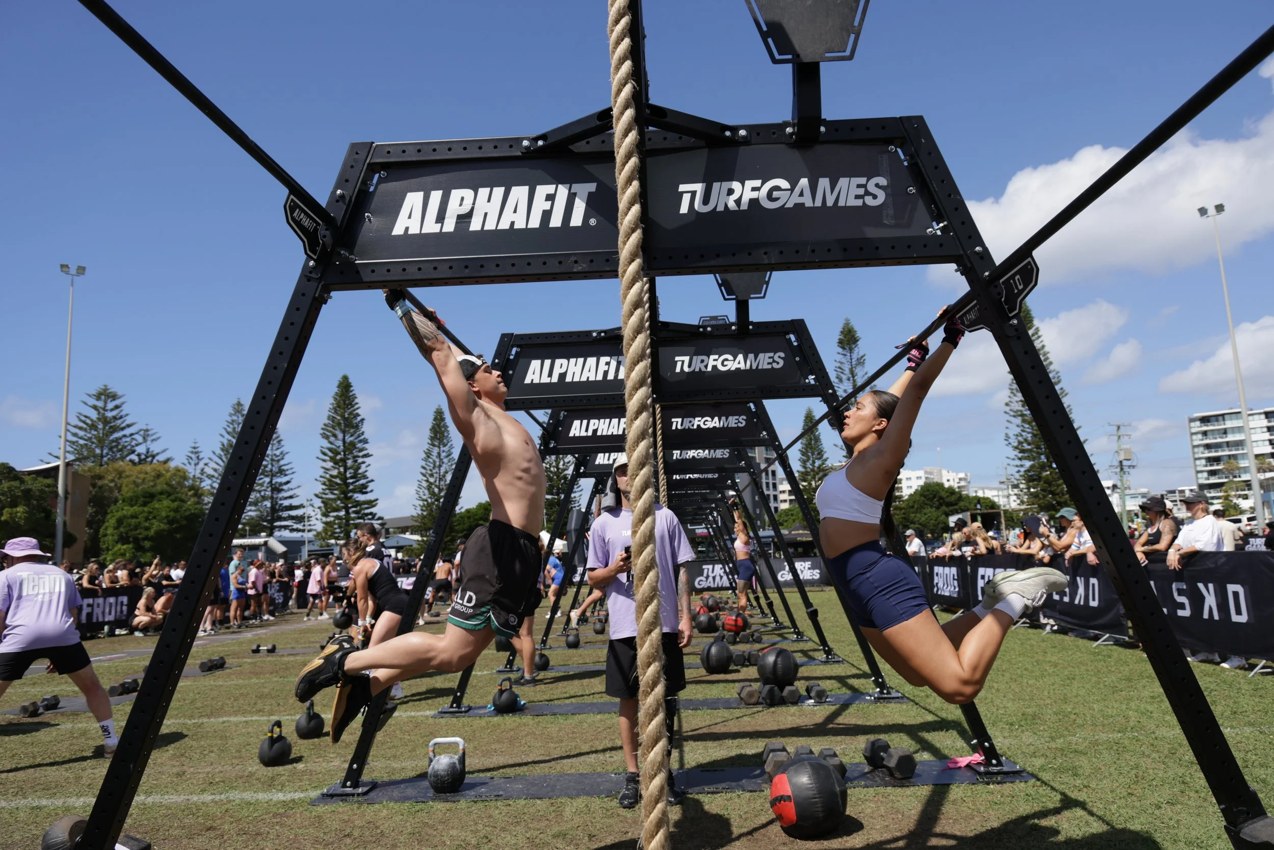 Two people, a man and a woman, are participating in an outdoor fitness challenge involving a climbing rope and a rig with weights and signage labeled 'ALPHAFIT' and 'TURFGAMES'. Many spectators and other participants are visible in the background on 