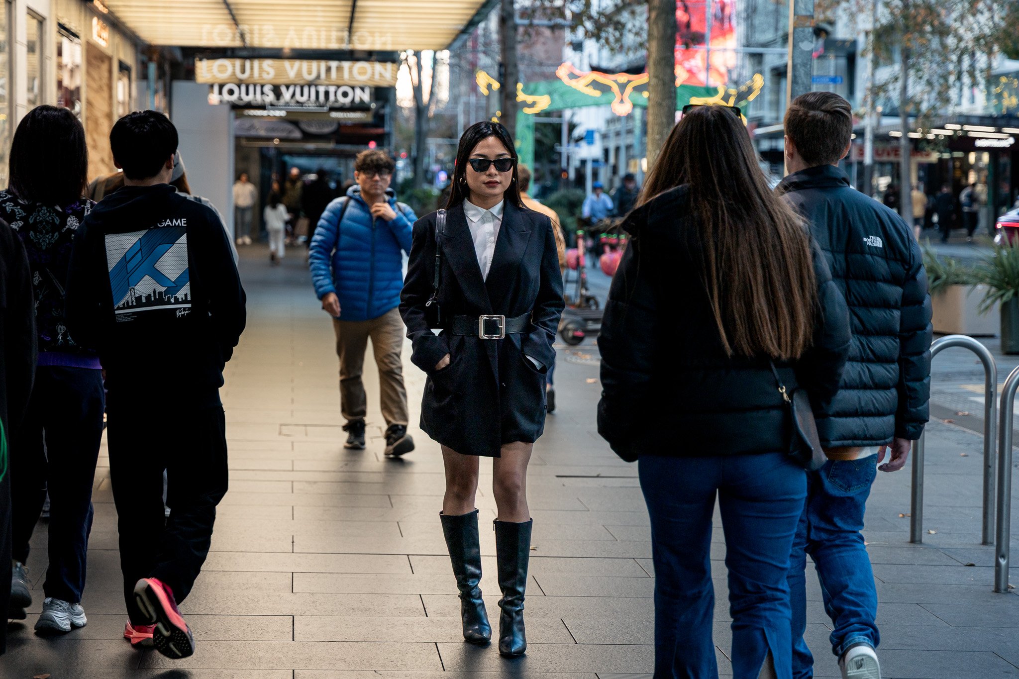 A woman walks confidently down a city sidewalk while other pedestrians are around her, with storefronts, signs, and trees in the background. The woman wears sunglasses, a dark blazer, a white shirt, a belt, and knee-high black boots.
