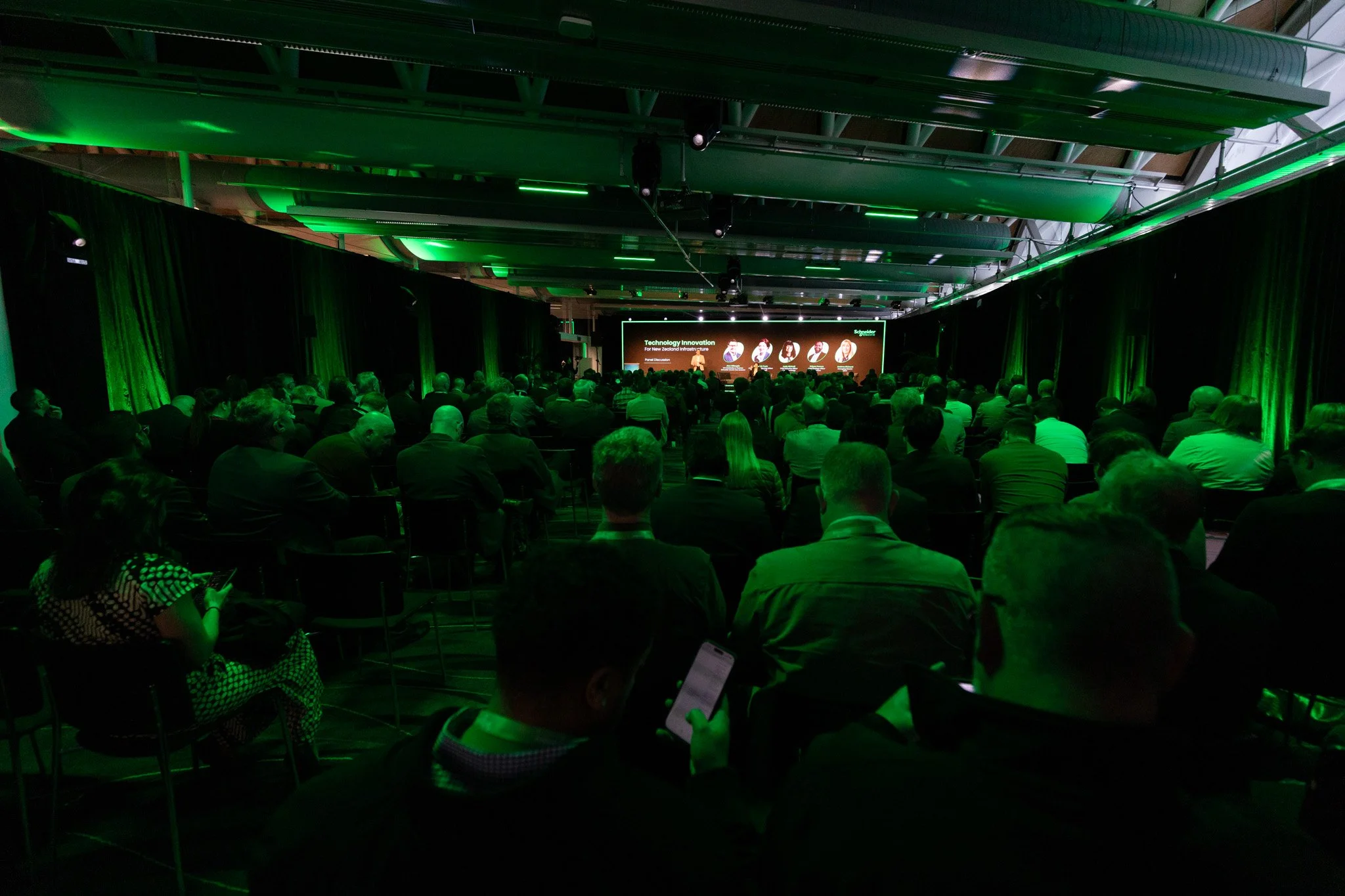 A large conference room filled with people attending a presentation. The room is dimly lit with green accent lighting and a large screen at the front displaying a slide titled 'Technology Innovation for New Zealand Infrastructure.'