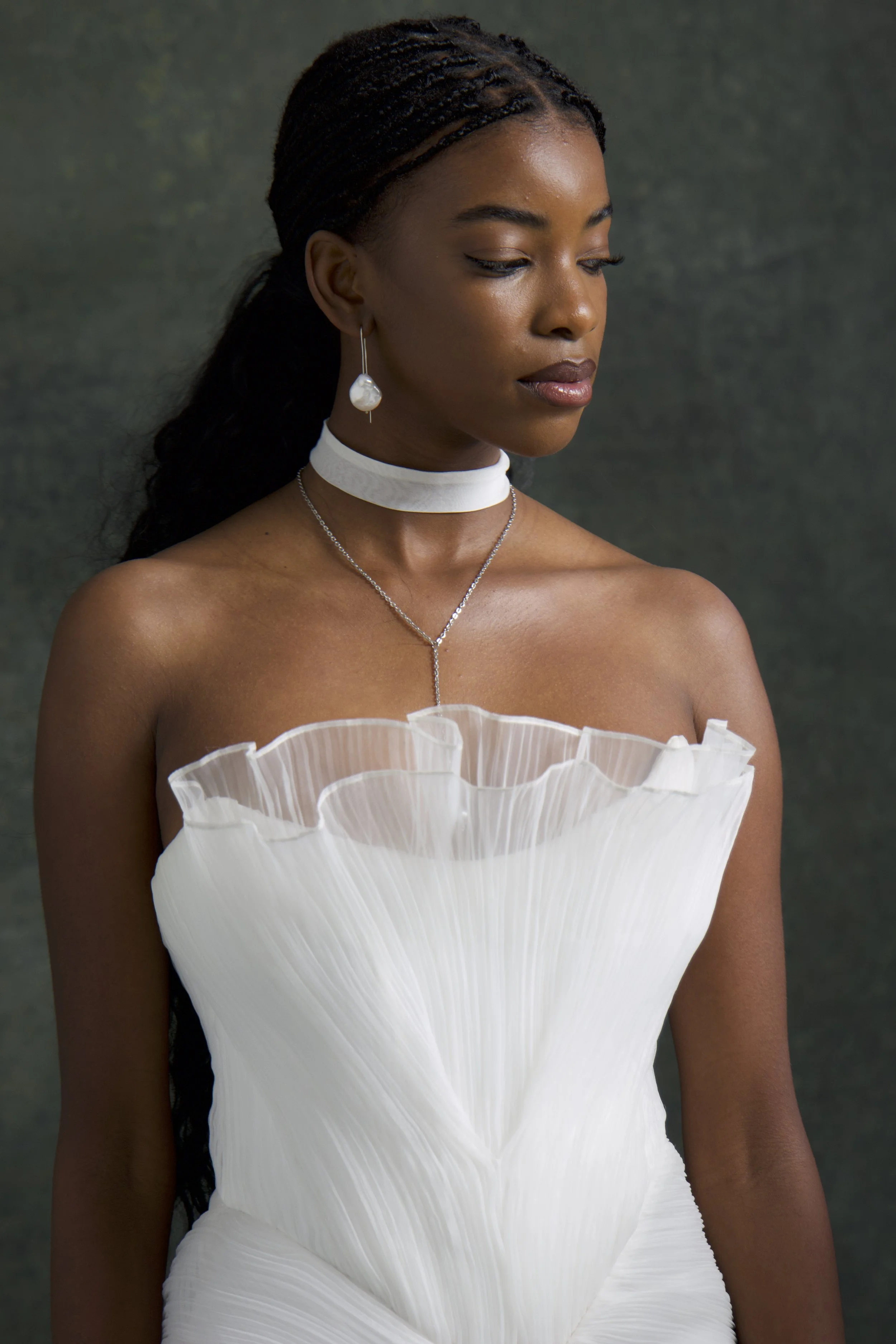 A woman with dark skin and braided hair, wearing a white strapless dress with ruffled details, a pearl earring, and layered necklaces, looking downward against a dark background.