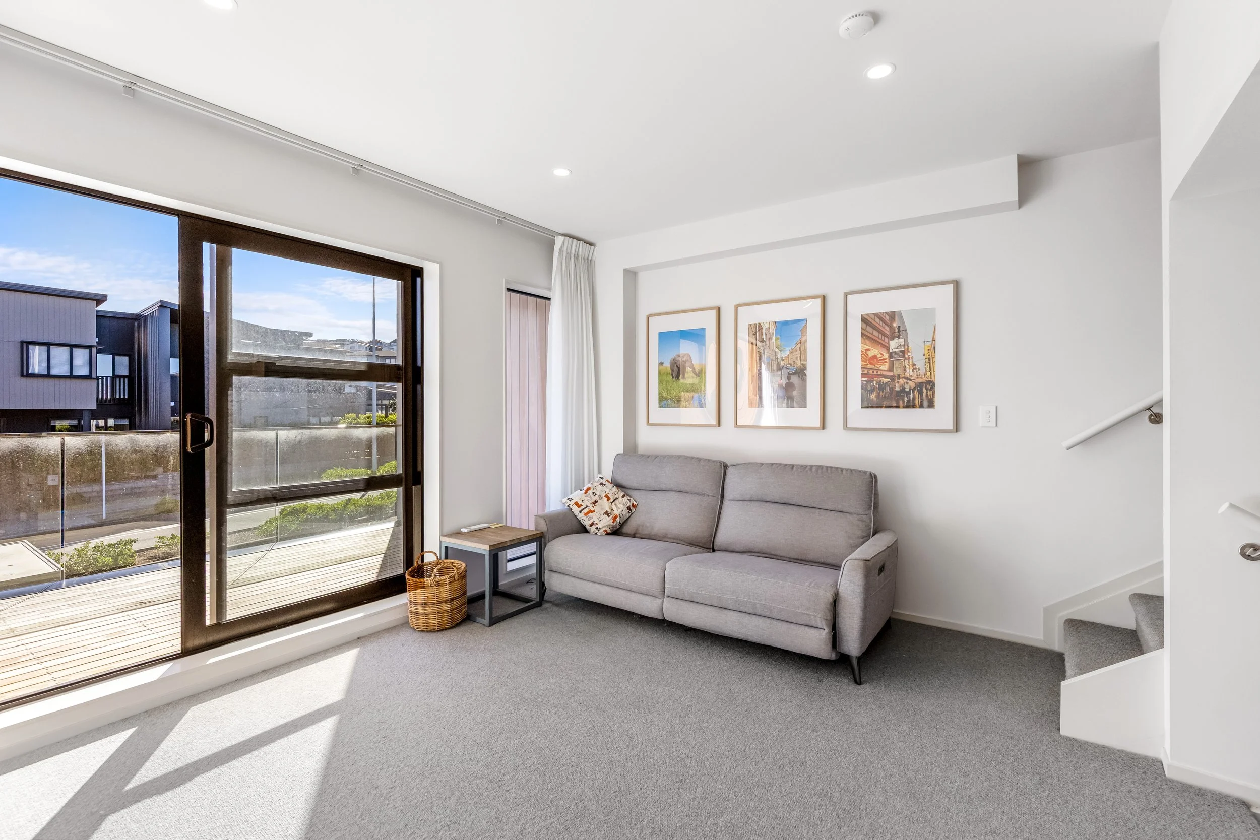 Living room with large sliding glass door leading to balcony, gray sofa with decorative pillows, framed photos on white wall, beige carpeted floor, and white ceiling with recessed lighting.