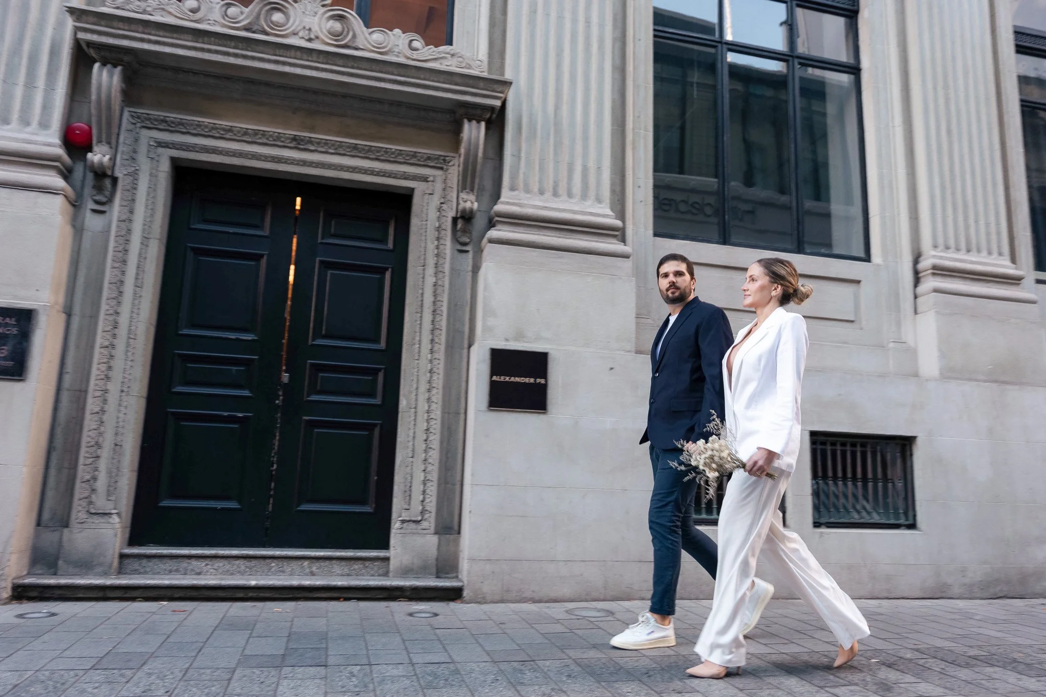 A man and woman dressed in formal attire walk together outside a historic building with grand columns and a large black door. The woman is carrying a bouquet of flowers.