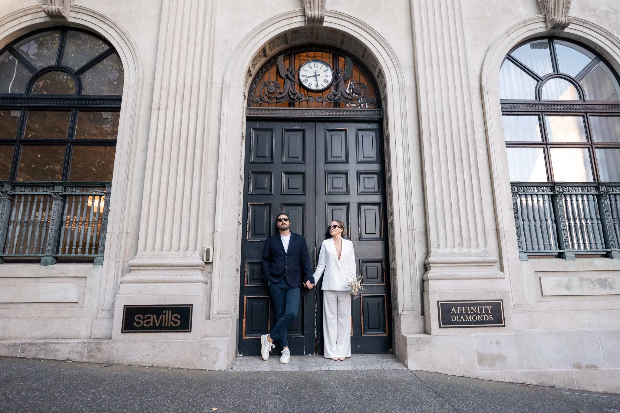 A man and woman in formal attire standing in front of a large, ornate building with a clock above the doorway. The man is wearing a dark suit and sunglasses, and the woman is wearing a white suit and sunglasses, holding a small bouquet of flowers.