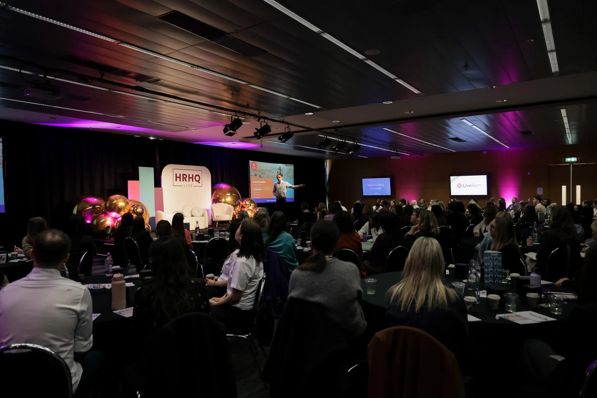 A large conference room filled with seated attendees listening to a speaker on stage. The stage has a sign that reads 'HRHQ LIVE' and several large decorative balloons. The room has dark ceiling panels with purple lighting accents and multiple screen