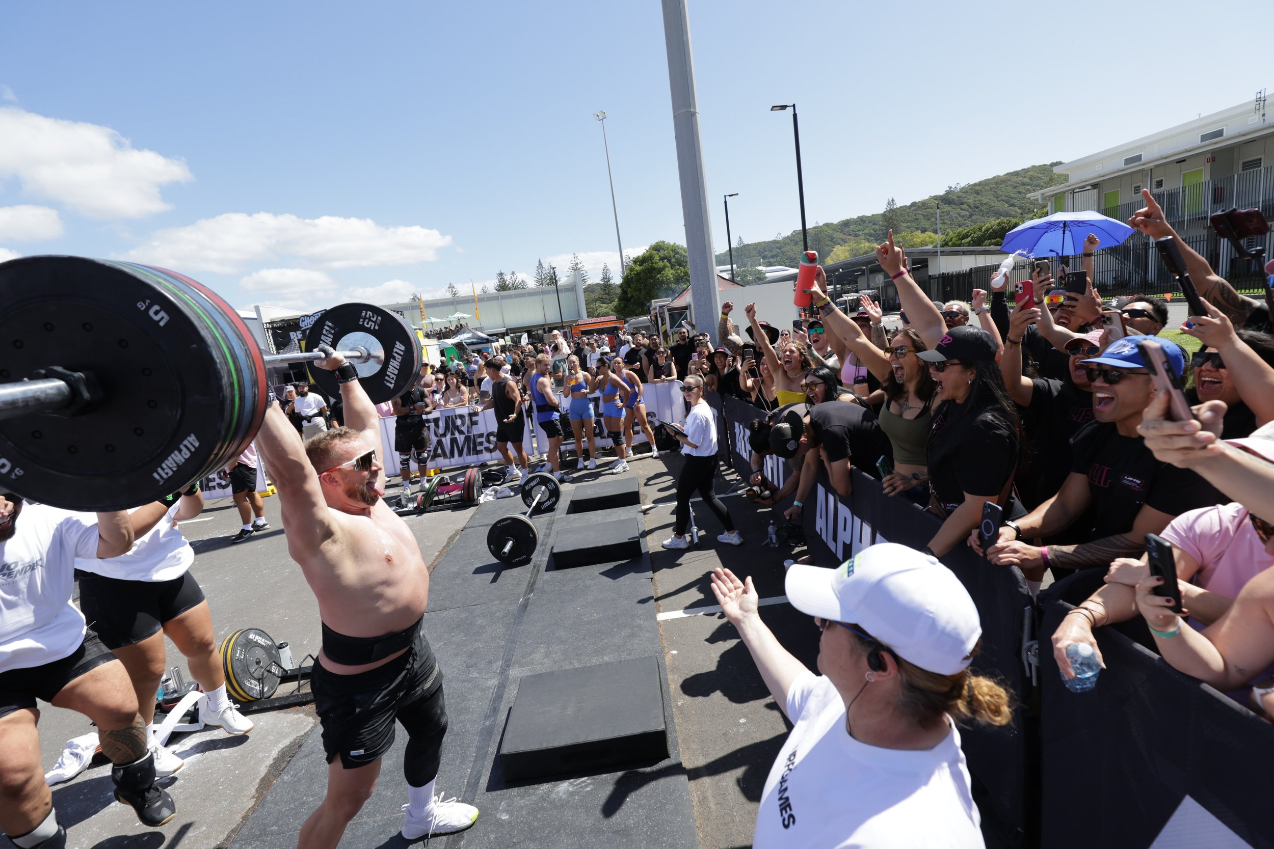 A muscular man performing a weightlifting exercise at an outdoor fitness event with a cheering crowd around him, some taking photos or videos, on a sunny day with a clear sky.