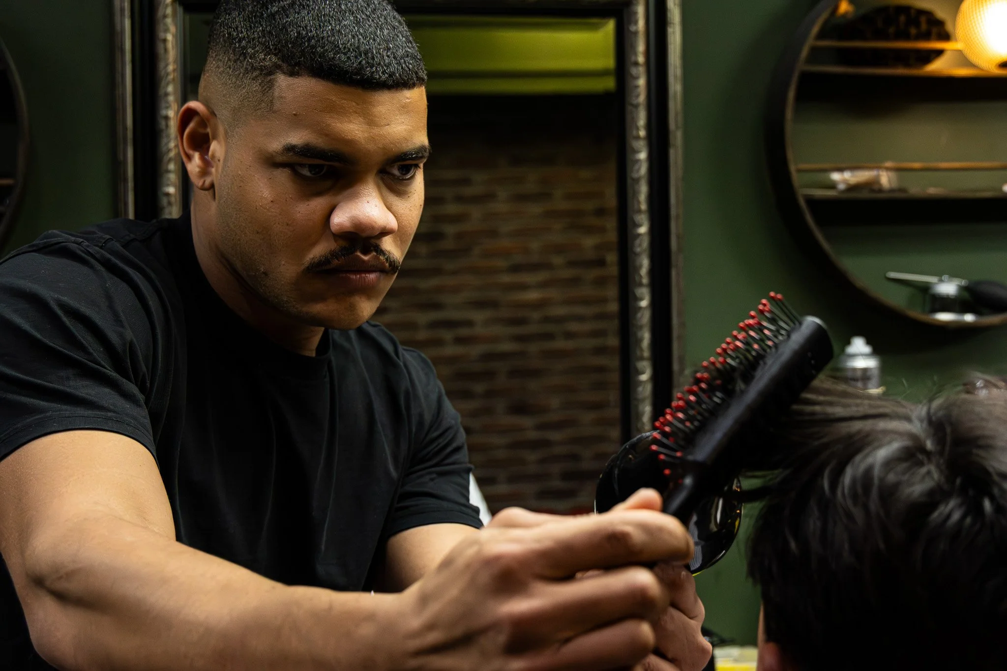 A man with short dark hair and a mustache, wearing a black shirt, is focused on grooming or cutting hair in a hair salon. He is holding a comb and scissors, working on a client's hair.