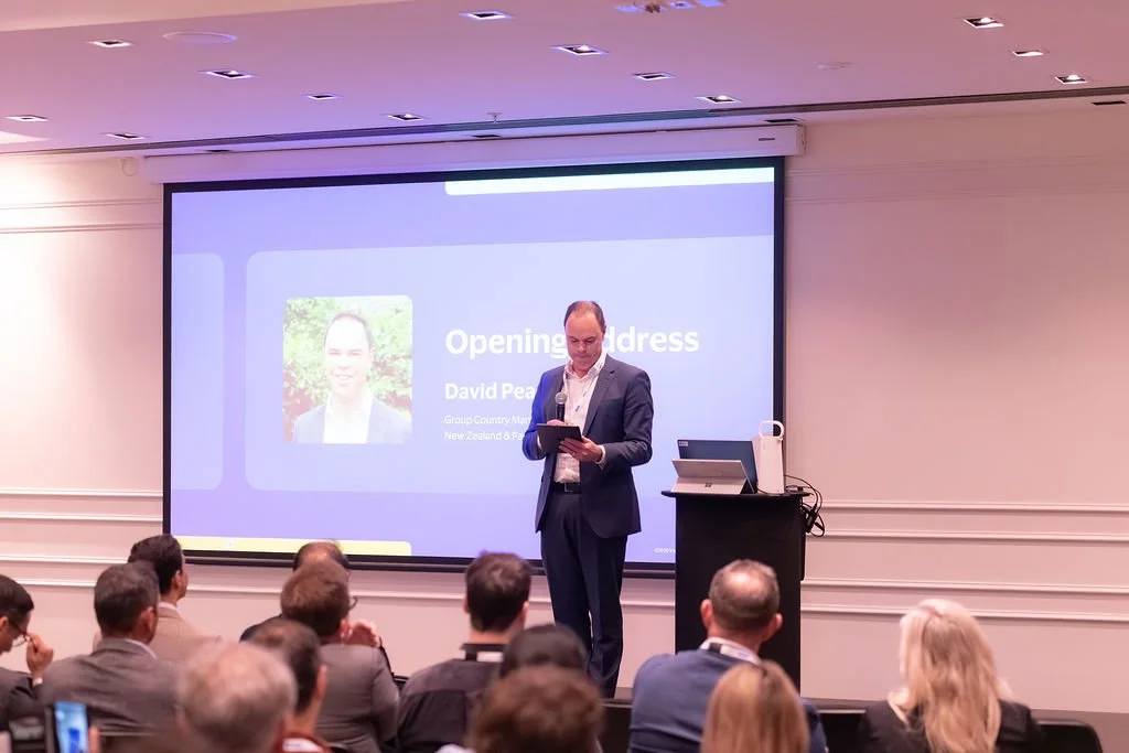 A man in a suit giving an opening address at a conference, standing in front of a screen displaying his name and title, with an audience seated in front of him.