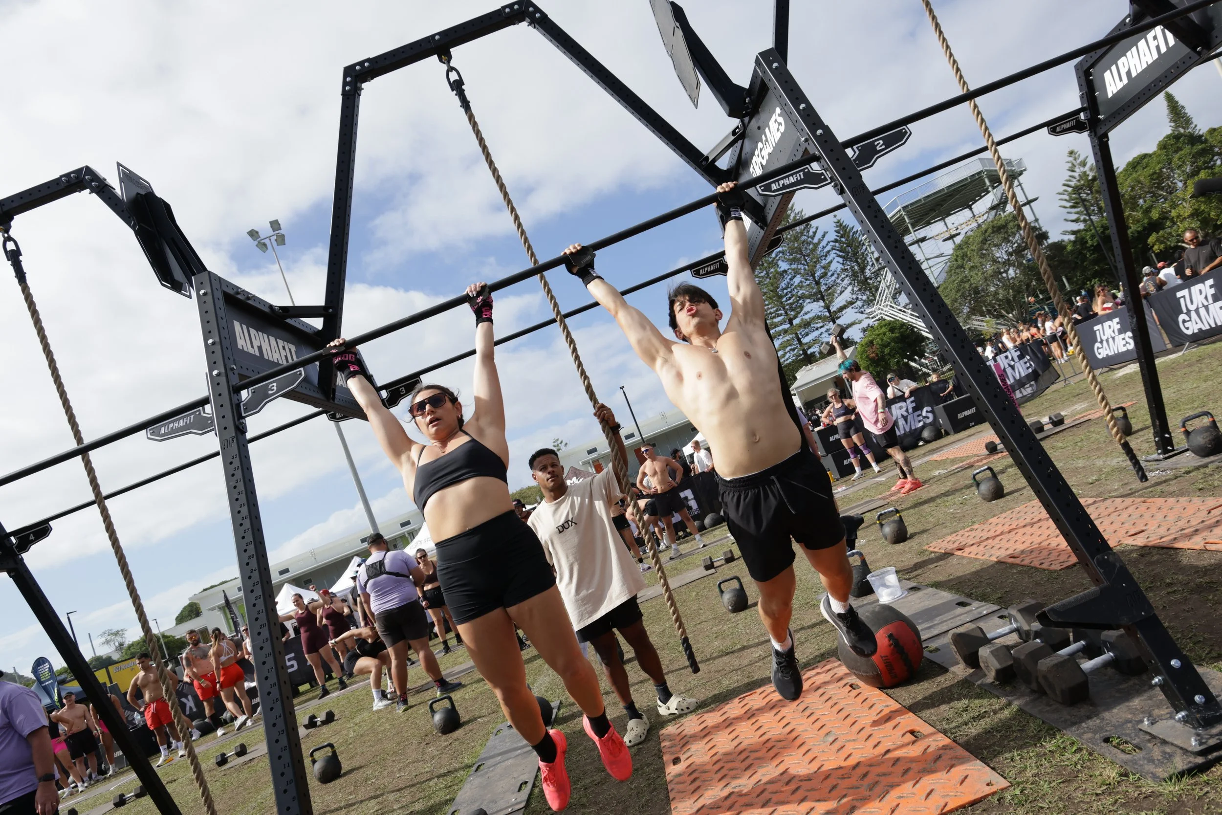 People participating in an outdoor fitness competition with black metal rigs, ropes, and weights, with spectators in the background on a sunny day.