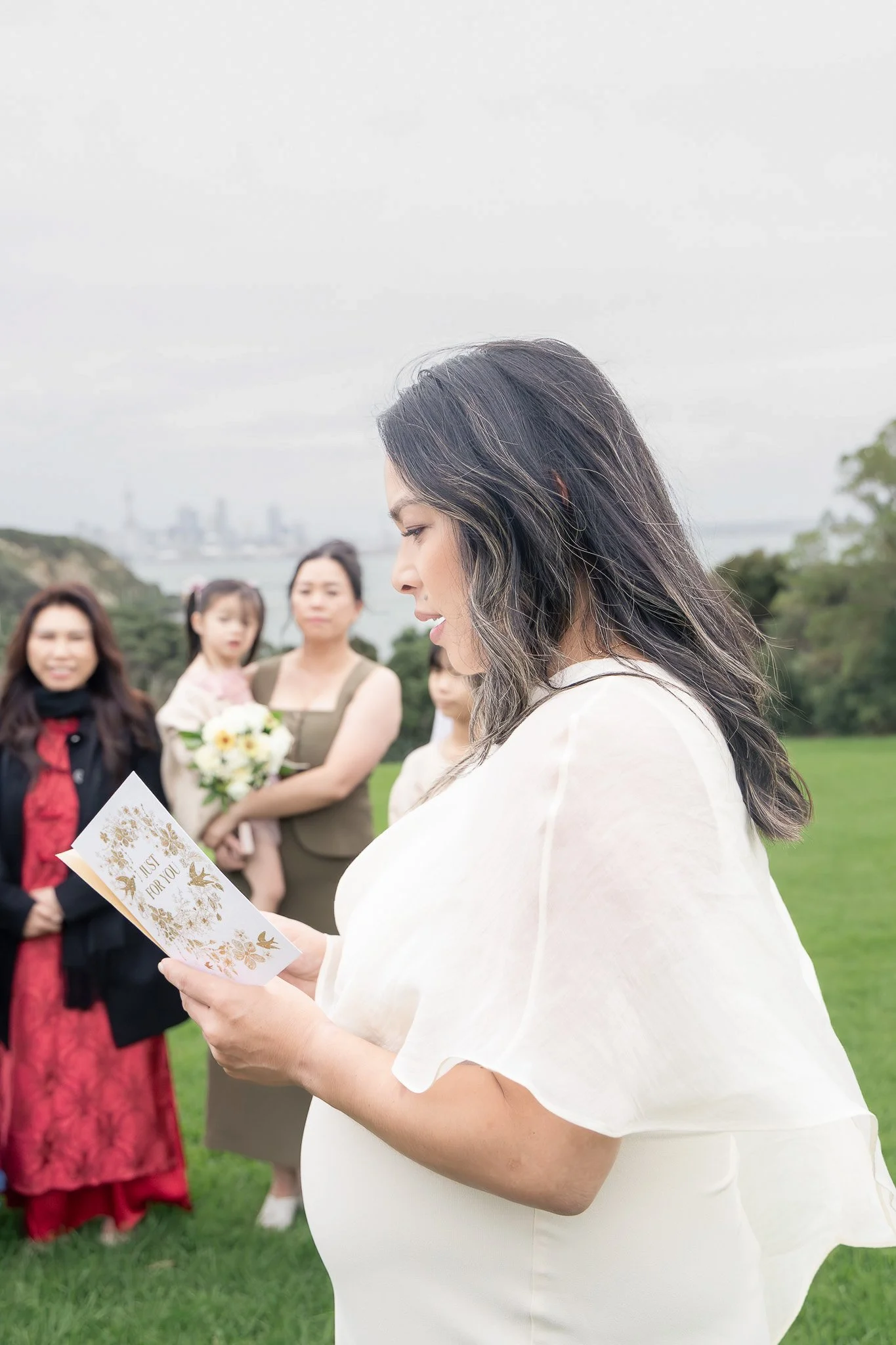 A pregnant woman with long dark hair reading a card during an outdoor gathering with several women, some in traditional dresses, on a grassy field with trees and a city skyline in the background.