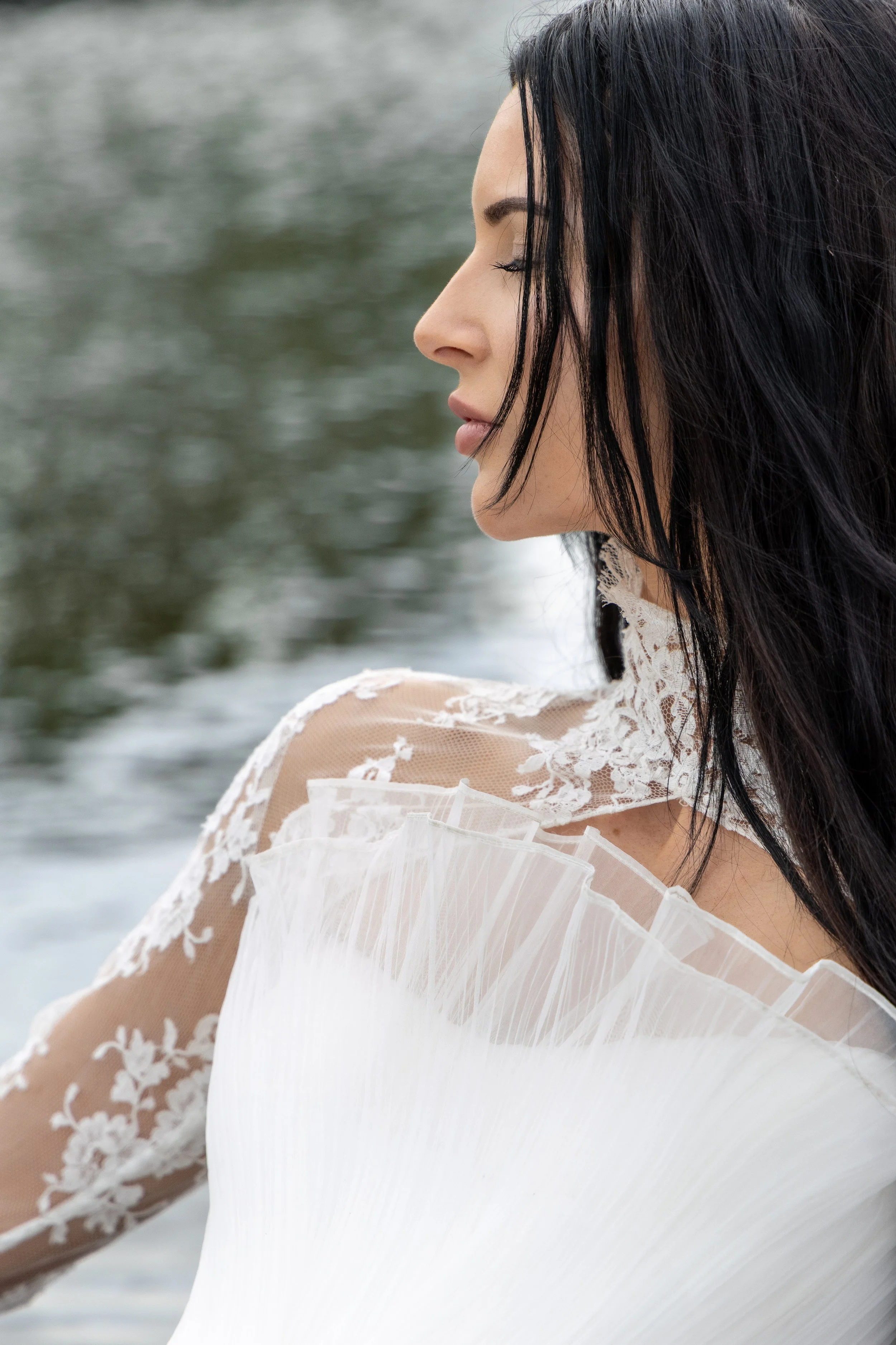 Side profile of a woman with long black hair, dressed in a white lace top, near water.