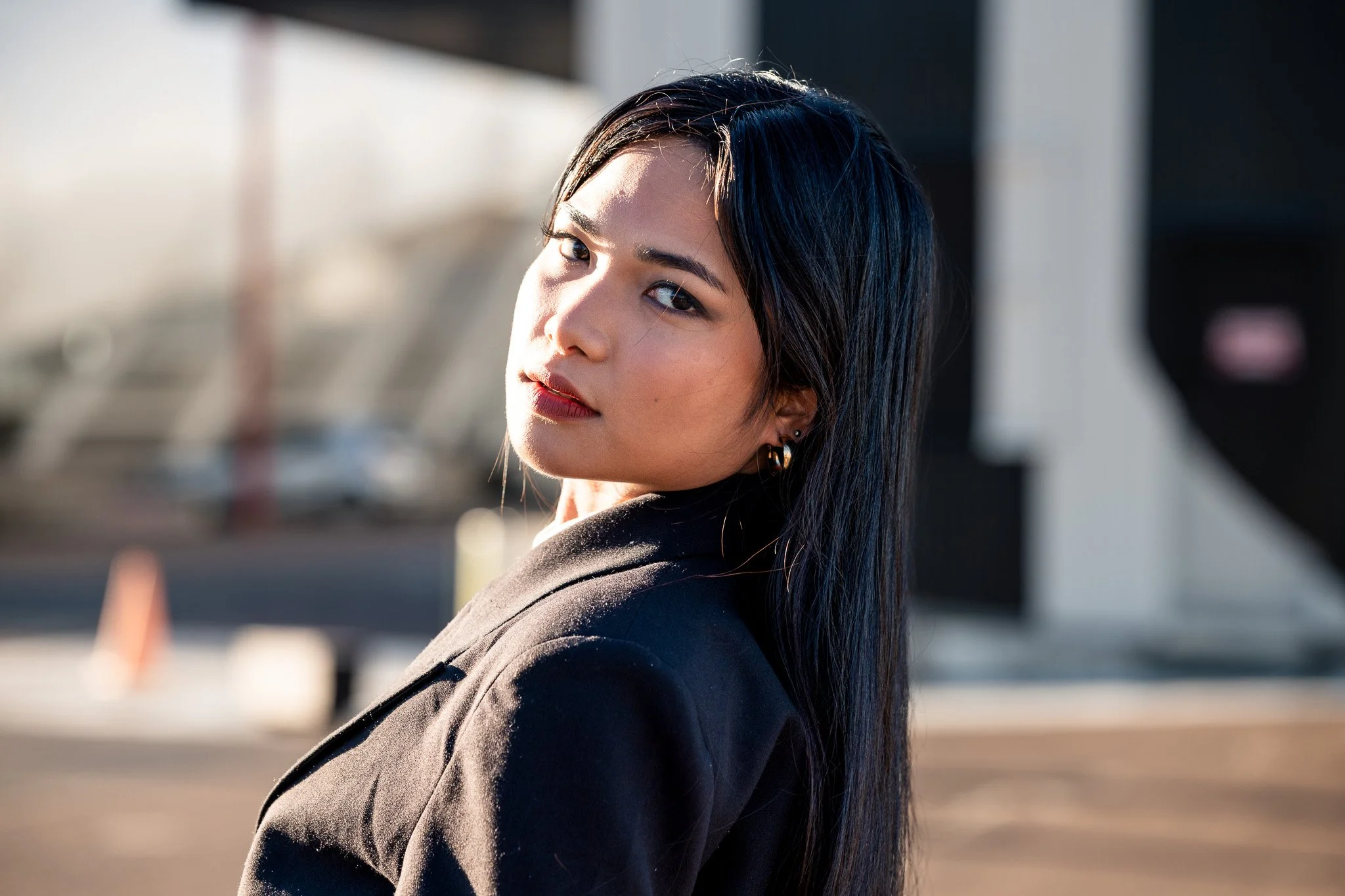 A young woman with dark hair and earrings looking over her shoulder in bright sunlight, wearing a black jacket.