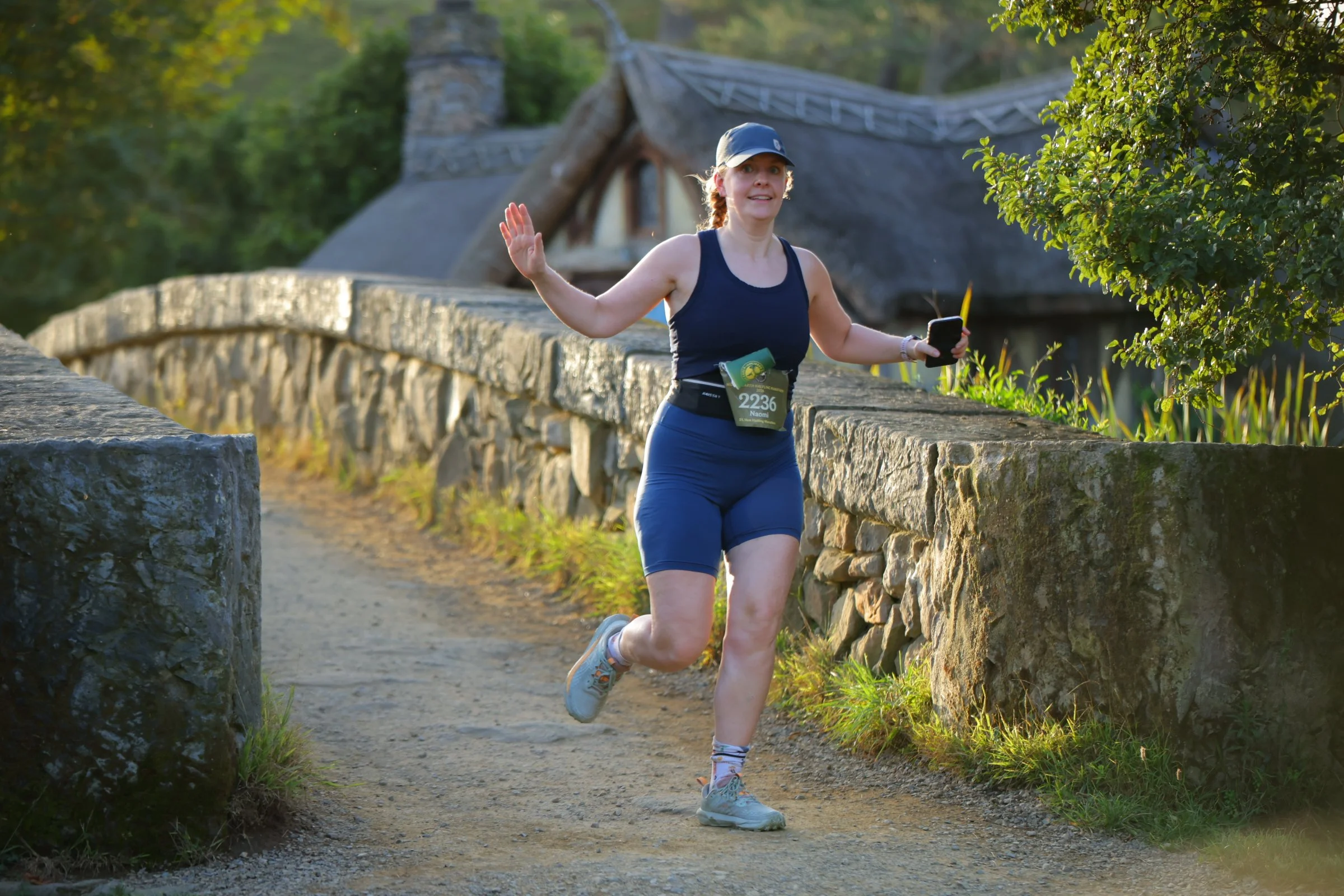 A woman jogging on a dirt path next to a stone wall, while holding a phone and raising her hand in a wave. She is wearing a blue tank top, blue shorts, a cap, and running shoes. There is a person with a race bib number 2236 on her waist, and the back