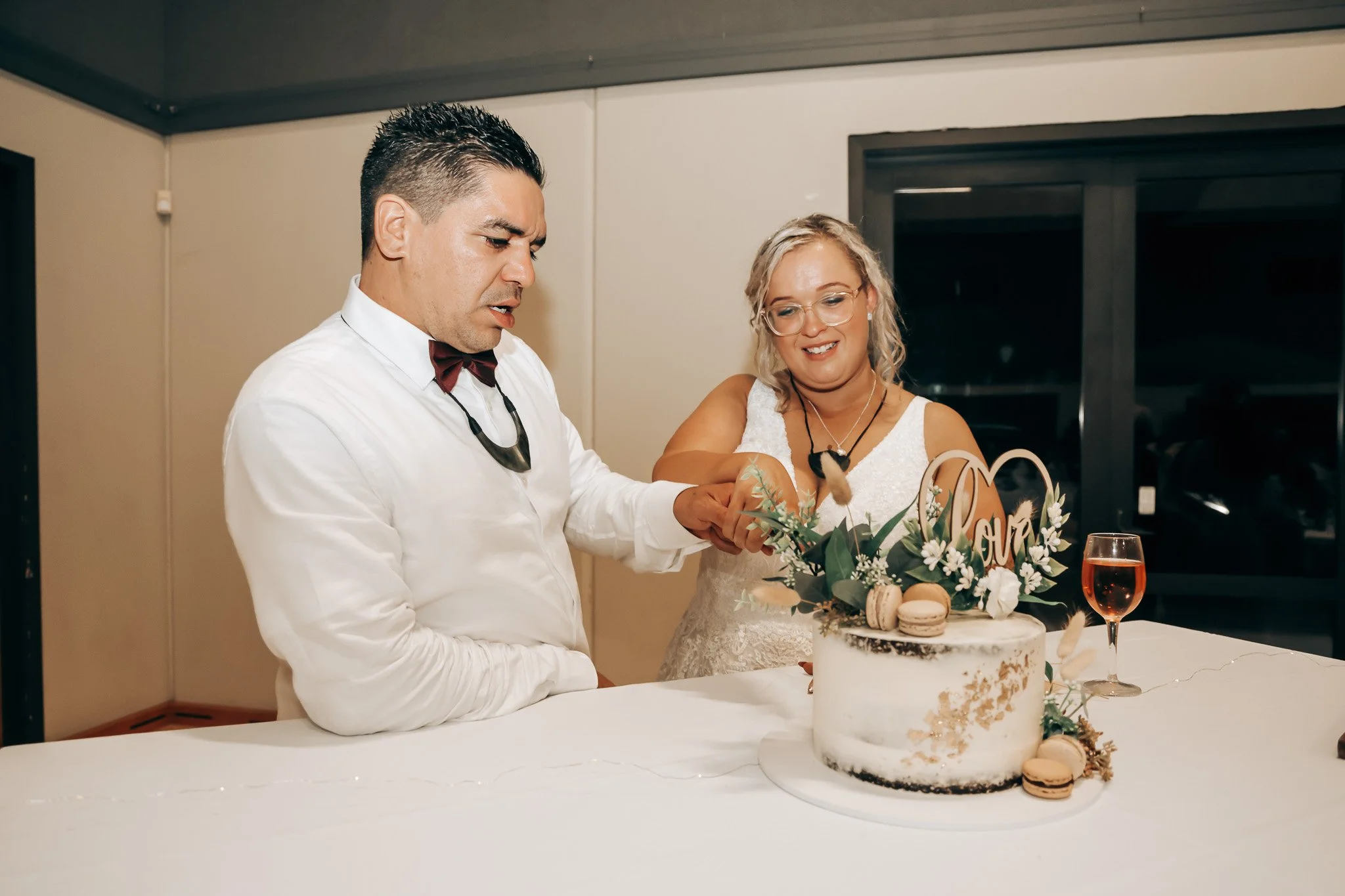 A man and woman at a celebration cutting a cake with macarons and a floral decoration, a glass of wine nearby, in an indoor setting.